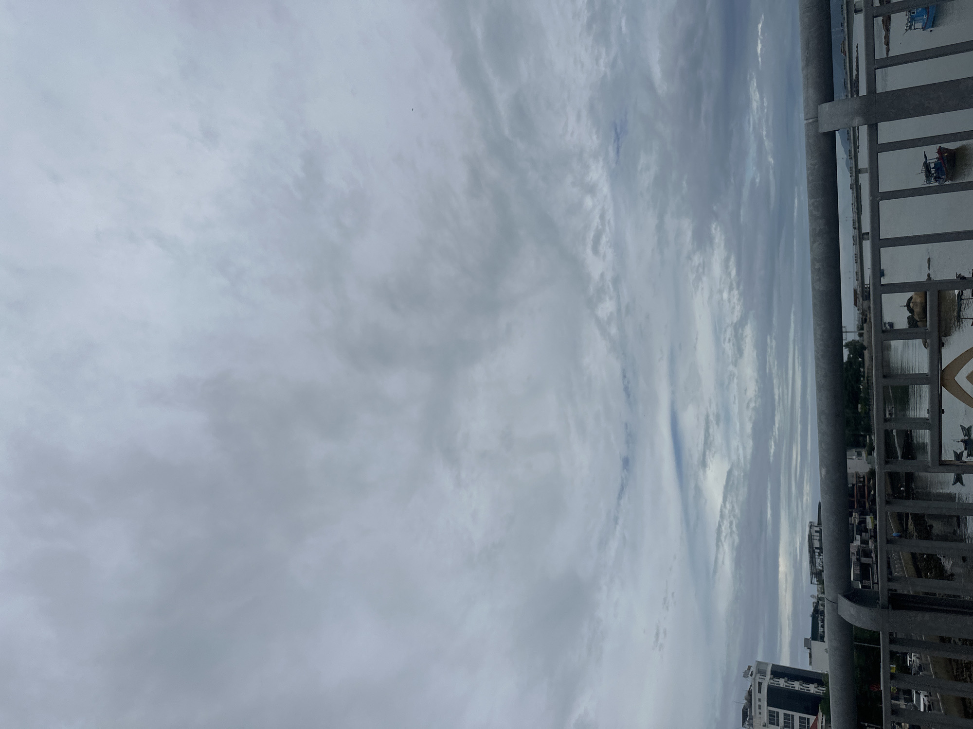 The image shows a view from a bridge in Bac Nha Trang Ward, Vietnam. The sky is overcast with high-altitude clouds. A metal railing runs vertically on the right side of the frame, overlooking the water and the cityscape. Several boats are visible on the water. Buildings are located in the background. The image is tilted or rotated.