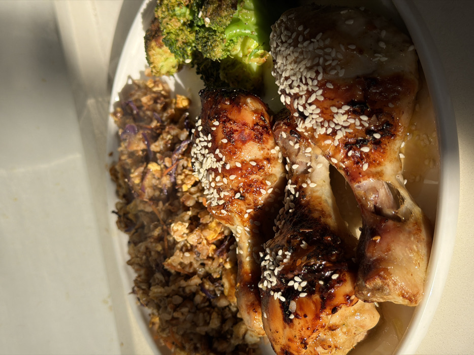 A close-up, high-angle image depicts a prepared meal served on a white, oval plate. The primary subject consists of three glazed chicken drumsticks, uniformly coated with white sesame seeds, positioned in the foreground. To the left and rear of the drumsticks is a mound of mixed grain or rice dish, appearing to contain shredded purple cabbage and other fine vegetable pieces. To the right of the drumsticks, several green florets of cooked broccoli are visible. The plate rests on a light-colored surface, with a light-colored vertical surface or wall in the background. Strong, directional natural light from the upper left illuminates the food, creating visible highlights on the glazed chicken and casting soft shadows. The scene is located in Tel-Aviv, Israel.