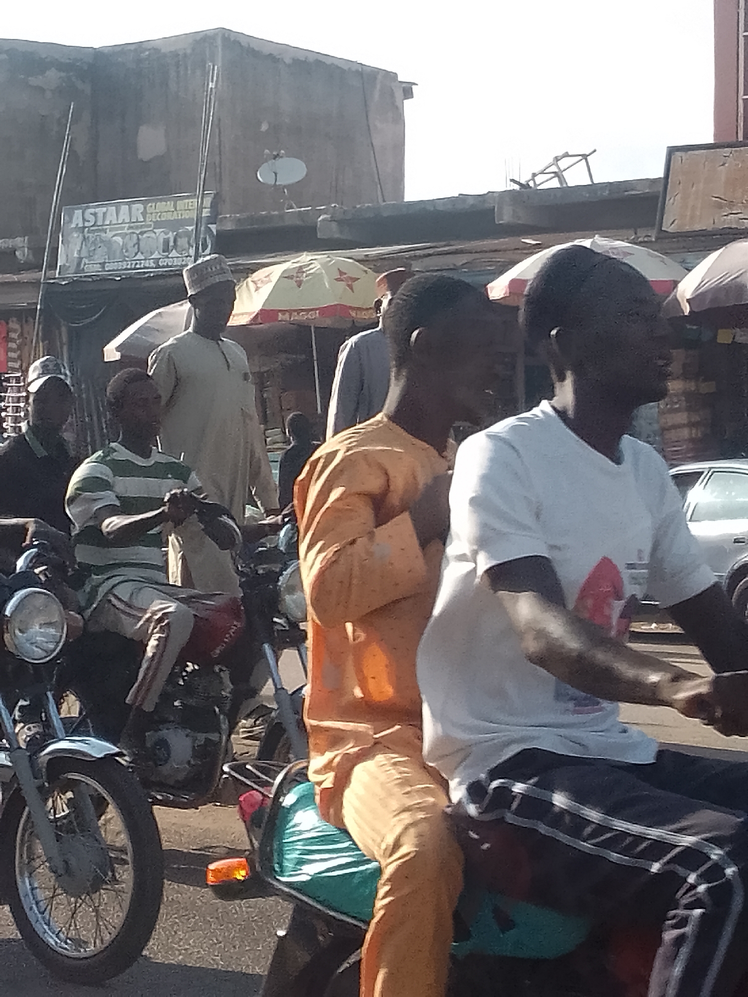 This image captures a bustling street scene in Arawa, Nigeria, under clear daylight conditions. In the immediate foreground, two males are positioned on a motorcycle; the passenger, facing left, wears an orange tunic and matching pants, while the driver behind him, also facing left, is clad in a white t-shirt and dark pants with white vertical stripes, seated on a seat with a teal cover. To their left, a male on a red motorcycle is visible, wearing a green and white striped t-shirt and light-colored pants, with his hands near the handlebars. Further left, a male in a dark shirt and a baseball cap is partially seen on another motorcycle.
In the mid-ground, a standing male in a light-colored tunic and a cap (kufi) is prominently featured. Behind these individuals, two market umbrellas are open: one yellow with a red "MAGGI" logo and another white. The background is dominated by a weathered, multi-story concrete building with a large signboard displaying "ASTAAR GLOBAL INTER DECORATION" and contact numbers "08033272745, 0703020". A small white satellite dish is mounted on the rooftop of this building. The foreground shows an asphalt road surface. Several other individuals are partially visible in the deeper background, contributing to the urban density.