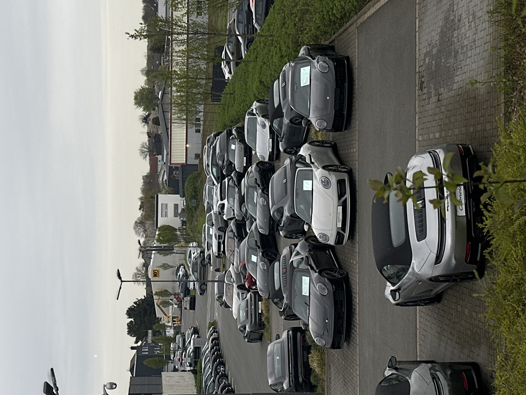 The image shows a car dealership in Holzwickede, Germany. The foreground shows a row of new Porsche vehicles parked on a paved lot, with some partially obscured by greenery. The cars are primarily gray, white, and black, with some models being convertibles. The vehicles appear to be ready for sale, as many have window stickers displayed. The dealership continues into the background, with numerous other cars parked in rows and on the street. Beyond the dealership are buildings, houses, and trees that make up the urban landscape of Holzwickede. The sky is overcast.