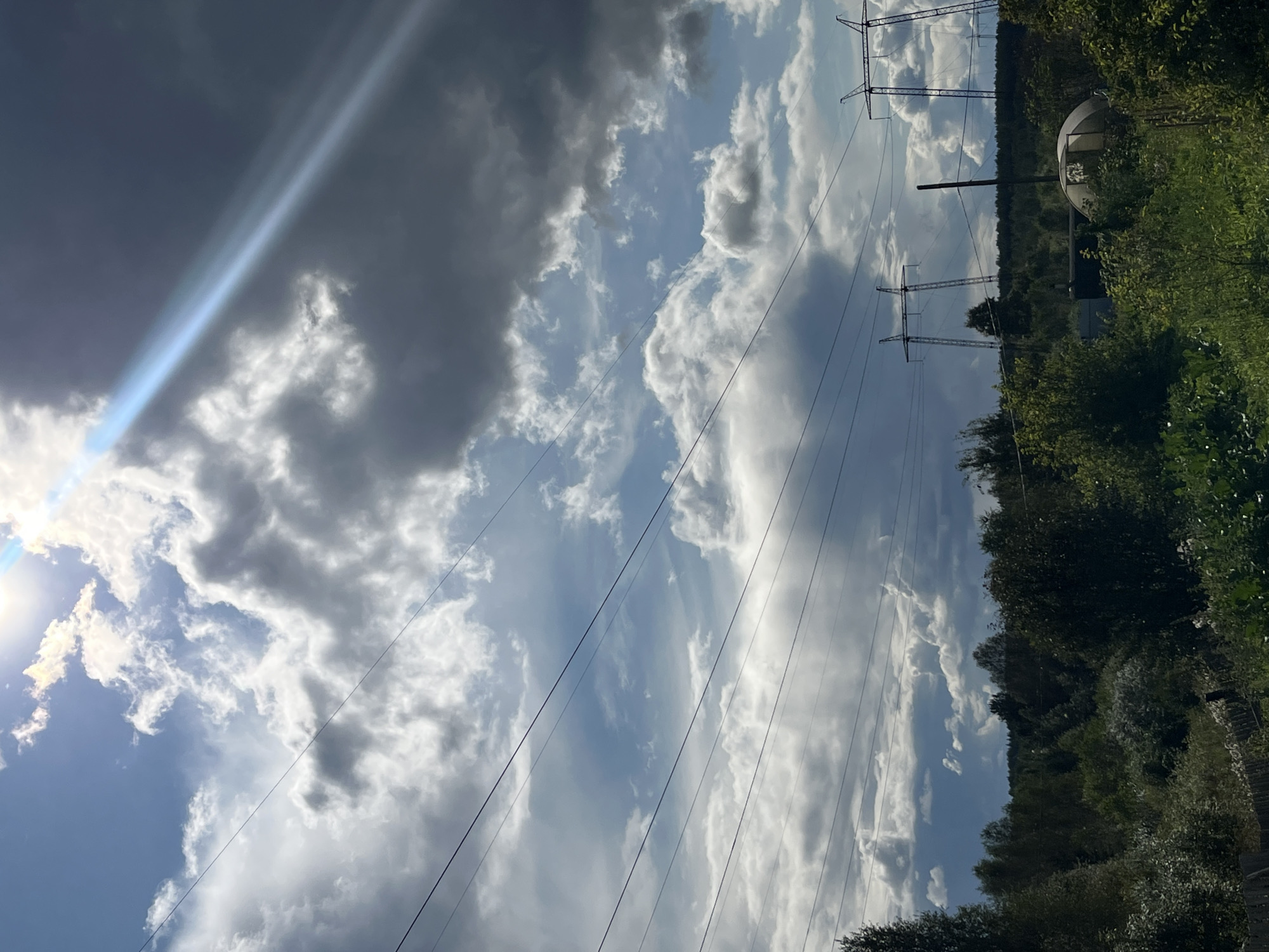 The image depicts a cloudy sky with a bright sunbeam cutting through the upper right portion. Several power lines stretch horizontally across the frame. Below the sky, a dense green treeline forms the horizon. In the foreground, there is lush vegetation, including bushes and trees. There are also structures visible in the distance, including what appears to be a greenhouse or dome-shaped structure and two large electrical towers. The overall impression is that of a rural or semi-rural landscape under a dramatic sky. The location is identified as Nizhny Tagil, Russia.