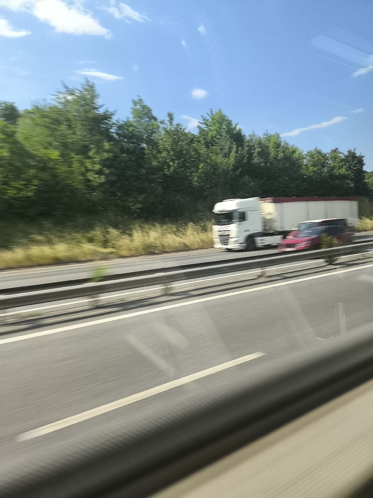 A blurred photograph taken from a moving vehicle on a highway shows a white semi-truck pulling a trailer, moving alongside a red car. The location is possibly near Saint-Chamond, France, given the likely European license plates and environmental characteristics. The highway has multiple lanes divided by white lines and guardrails. Trees and vegetation are visible along the roadside. The sky is blue with scattered clouds. The photograph's motion blur indicates a high speed of the vehicles.