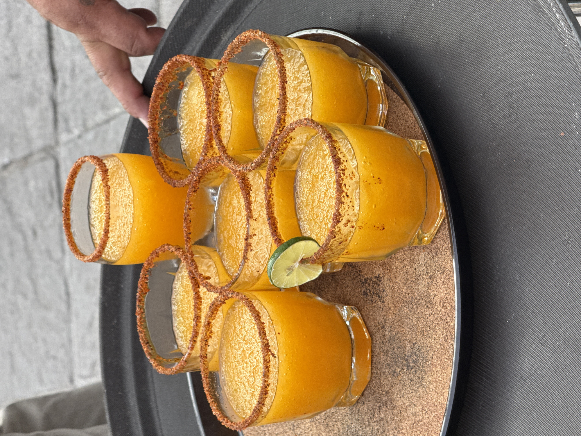 The image shows a tray of seven identical mango-colored beverages in glasses, each rimmed with a dark seasoning, possibly chili powder. A slice of lime garnish is placed on one of the glasses. The drinks are served on a round, textured tray with a black rim, held by a person whose hand is visible on the upper left. The scene takes place in Santiago de Querétaro, Mexico.