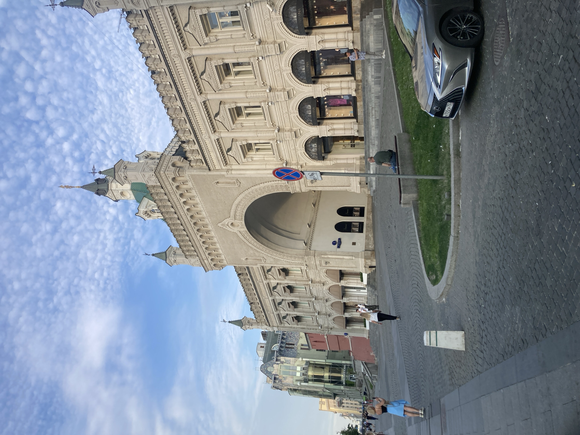 The image captures a street scene in Moscow, Russia, likely near Red Square or the Kremlin, given the architectural style of the building dominating the frame. The main structure is a large, ornate building with a grand arched entrance and classical architectural elements, featuring multiple windows and decorative stonework. In the foreground, a cobblestone street is visible with a parked silver Lexus sedan. Several people are present: a woman in a blue dress is taking a photo with her phone on the left side of the street, while another woman in black and white clothing walks away from the camera. A man in a green shirt sits on a bench near the street, and another man in a patterned shirt stands to the right of the arched entrance. The sky above is blue with scattered white clouds, indicating daytime. The background shows a line of buildings, some with modern facades and others with more traditional designs.