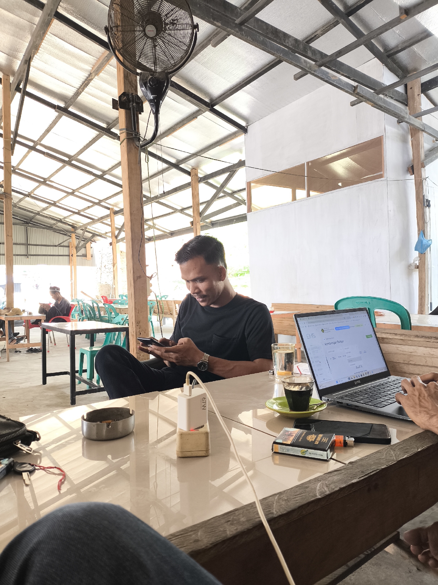In Keudejungkagajah, Indonesia, a man wearing a black t-shirt and dark pants is seated at a table, looking down at his smartphone and interacting with it. He has a watch on his left wrist. On the table in front of him is a laptop displaying a webpage with text and images, a glass of water, a small cup of coffee on a green saucer, a pack of cigarettes, a lighter, and a phone charger plugged into an outlet. Behind him, other individuals are seated at tables in an open-air structure with a corrugated metal roof supported by wooden beams and poles. The environment appears to be a casual outdoor cafe or gathering place.