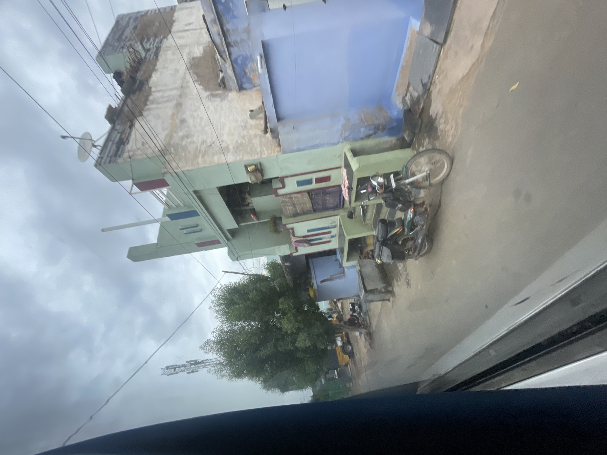 The image depicts a street scene in Bethamcherla, India, under an overcast sky. A multi-story building, partially painted in light green and off-white, dominates the right side of the frame. The building has a satellite dish on its roof and appears to be in a state of disrepair in some sections. Adjacent to it is another building with a bright blue facade. In the foreground, a green and black motorcycle is parked on the paved street. Further down the street, a small yellow vehicle and trees are visible. There are also several utility poles and wires crisscrossing the sky. The overall impression is of a typical street in a semi-urban or rural Indian setting.