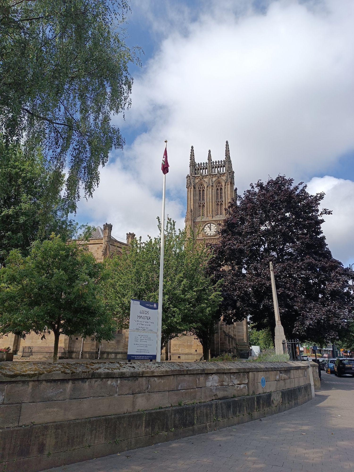 The image depicts Leeds Minster, also known as the Parish Church of St Peter, located in Leeds, United Kingdom. The view is from across a cobbled pavement, with a stone wall in the foreground. Behind the wall, trees partially obscure the view of the church's stone facade. To the left, a large deciduous tree with light green leaves is visible, while to the right, a tree with dark purple leaves is more prominent. The church's tower, a focal point of the image, features gothic architectural elements, including spires and arched windows, with a clock face visible. A flag flies from a flagpole in front of the tower. A sign identifying "Leeds Minster" and providing information about services is positioned to the left of the flagpole. A war memorial obelisk stands to the right of the purple-leaved tree. Several cars are parked and driving on a road visible in the background, with shops and buildings lining the street. The sky is partly cloudy, with patches of blue sky and white clouds. The scene appears to be set during the daytime, under natural light.