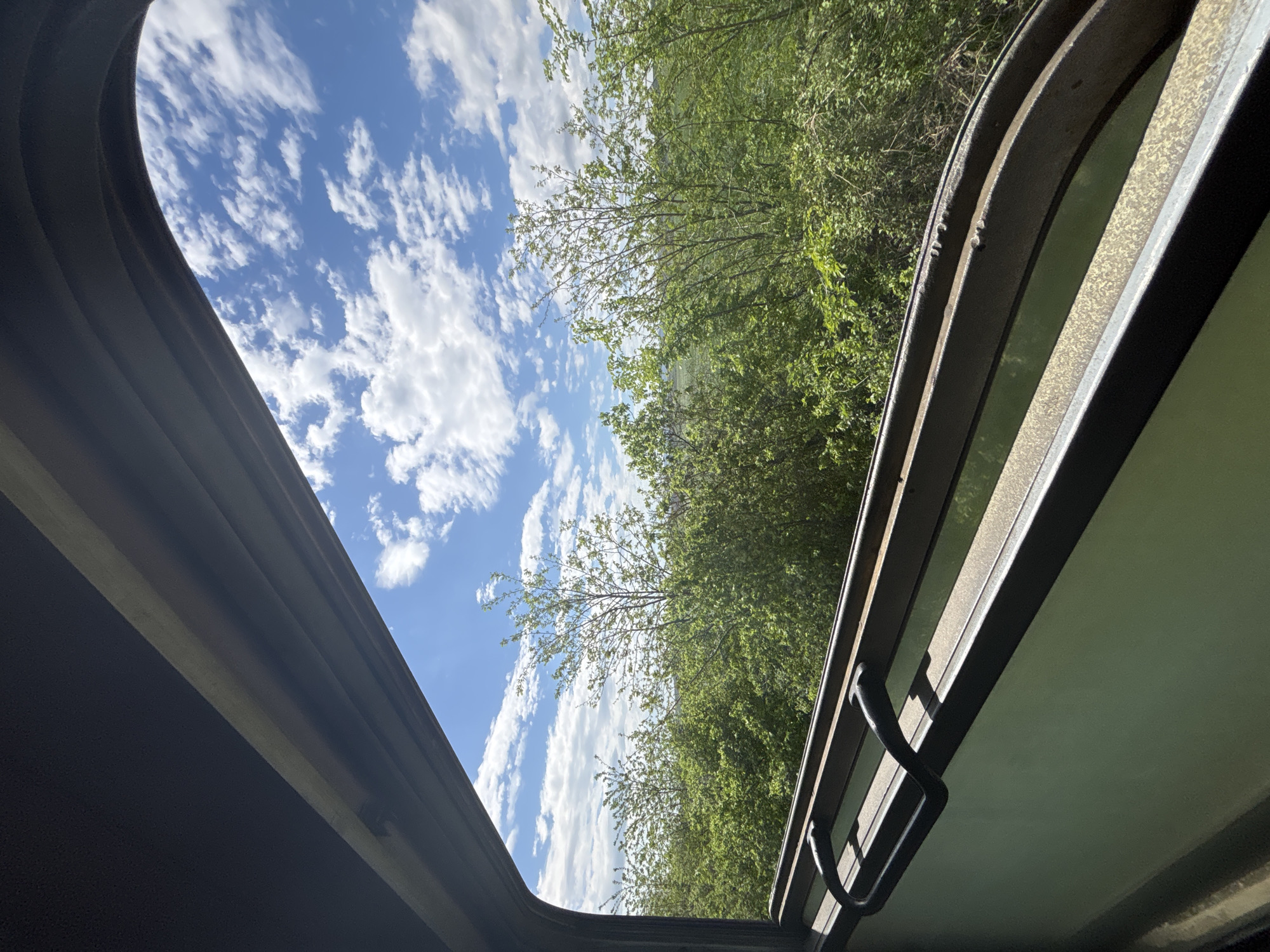 An open sunroof offers a view of a blue sky with scattered white clouds and the green foliage of trees. The sunroof's frame is visible on both sides, made of metal.