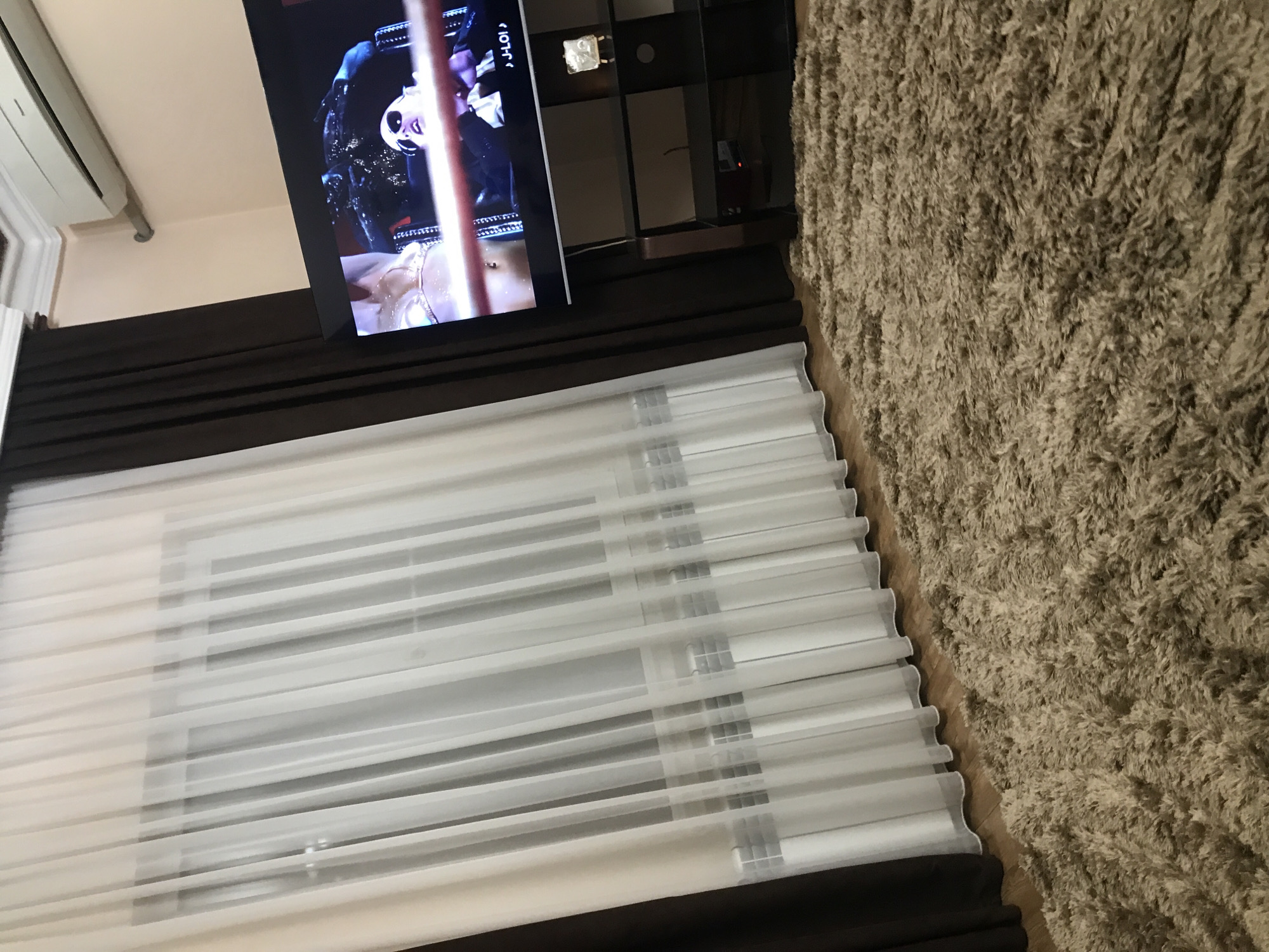 The image shows a living room with a TV mounted on the wall, displaying an image of a person performing on a pole. Below the TV, there is a window covered with white blinds and dark brown curtains. To the right of the window, there is a large, shaggy brown rug. The location is potentially in Yerevan, Armenia, based on the prompt but there is no element in the photo to confirm it.