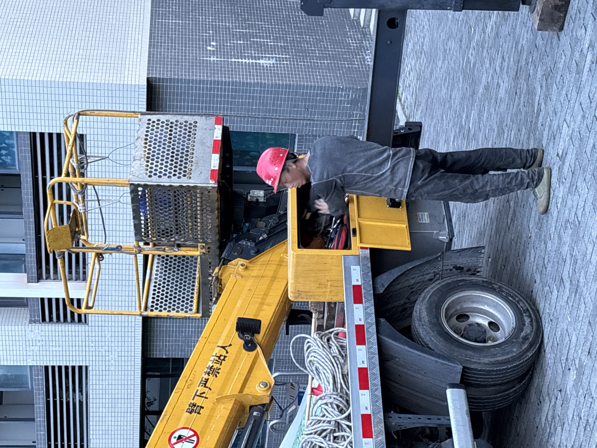 A person wearing a red hard hat and dark clothing is working on the machinery of a hydraulic platform lift in an outdoor setting, possibly a construction or maintenance site. The platform lift is yellow and black. A set of white ropes is bundled on the side of the truck. The background includes a building with gray and white tiled exterior walls. The photo was taken in the city of Jiangjin, China.