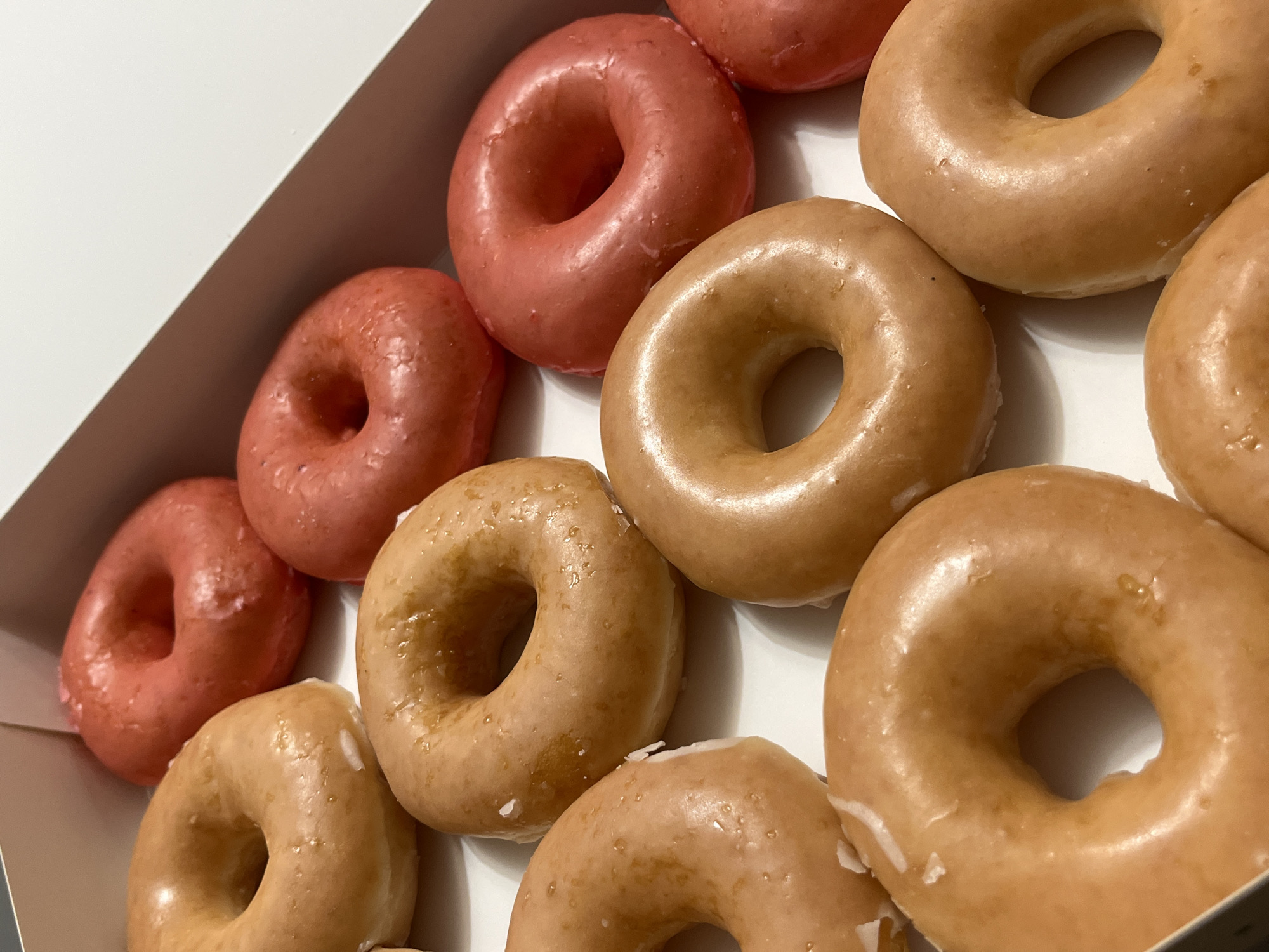 A box of doughnuts, with a mix of glazed and pink-frosted doughnuts, sits on a white surface in Unknown, Unknown. The doughnuts are arranged in rows within the box. The glazed doughnuts have a light brown, shiny coating. The pink doughnuts have a similar shiny surface with a light pink hue.