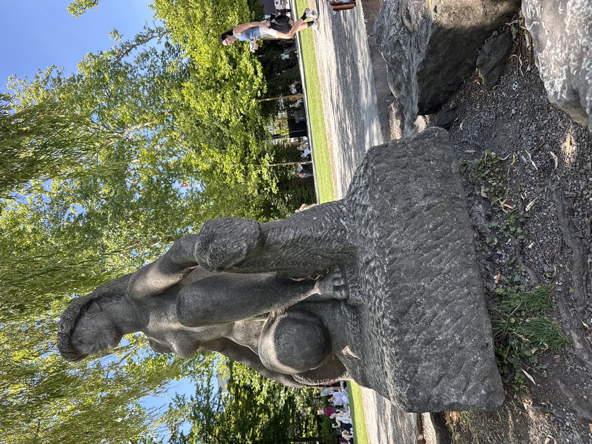 In the city of Lucerne, Switzerland, an outdoor scene unfolds with a stone sculpture of a woman as the primary subject. The sculpture is positioned in the foreground, resting on a rough stone base. Behind the sculpture, a park setting is visible, characterized by lush green trees and a clear blue sky. Several individuals are scattered throughout the park. In the middle ground, a woman walks from right to left, holding a drink in her right hand and a bag in her left. Further in the background, groups of people are seated on a grassy area. The ground surrounding the sculpture is a mixture of gravel and dirt, with some exposed tree roots. The overall impression is a serene day in a public park.