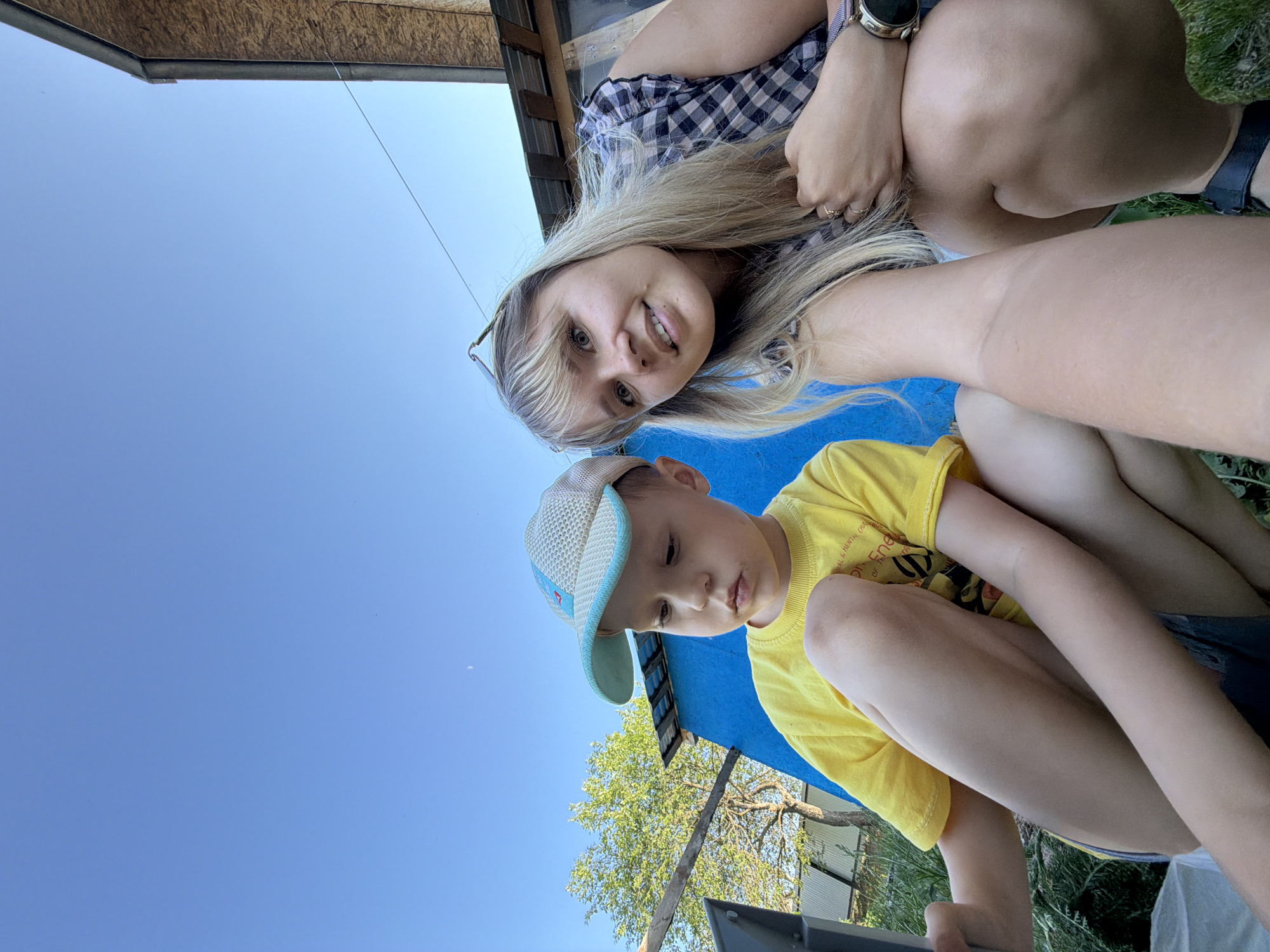The image depicts a woman and a child in an outdoor setting. The woman, with blonde hair, is wearing a checkered shirt and a watch on her left wrist. The child is wearing a yellow shirt and a blue and white cap. They are both in a crouching position, looking down. In the background, there is a blue tarp, some greenery, a tree, and a building with a wooden facade. The sky is clear blue. The exact location in Чумна, Russia cannot be determined from the image alone.