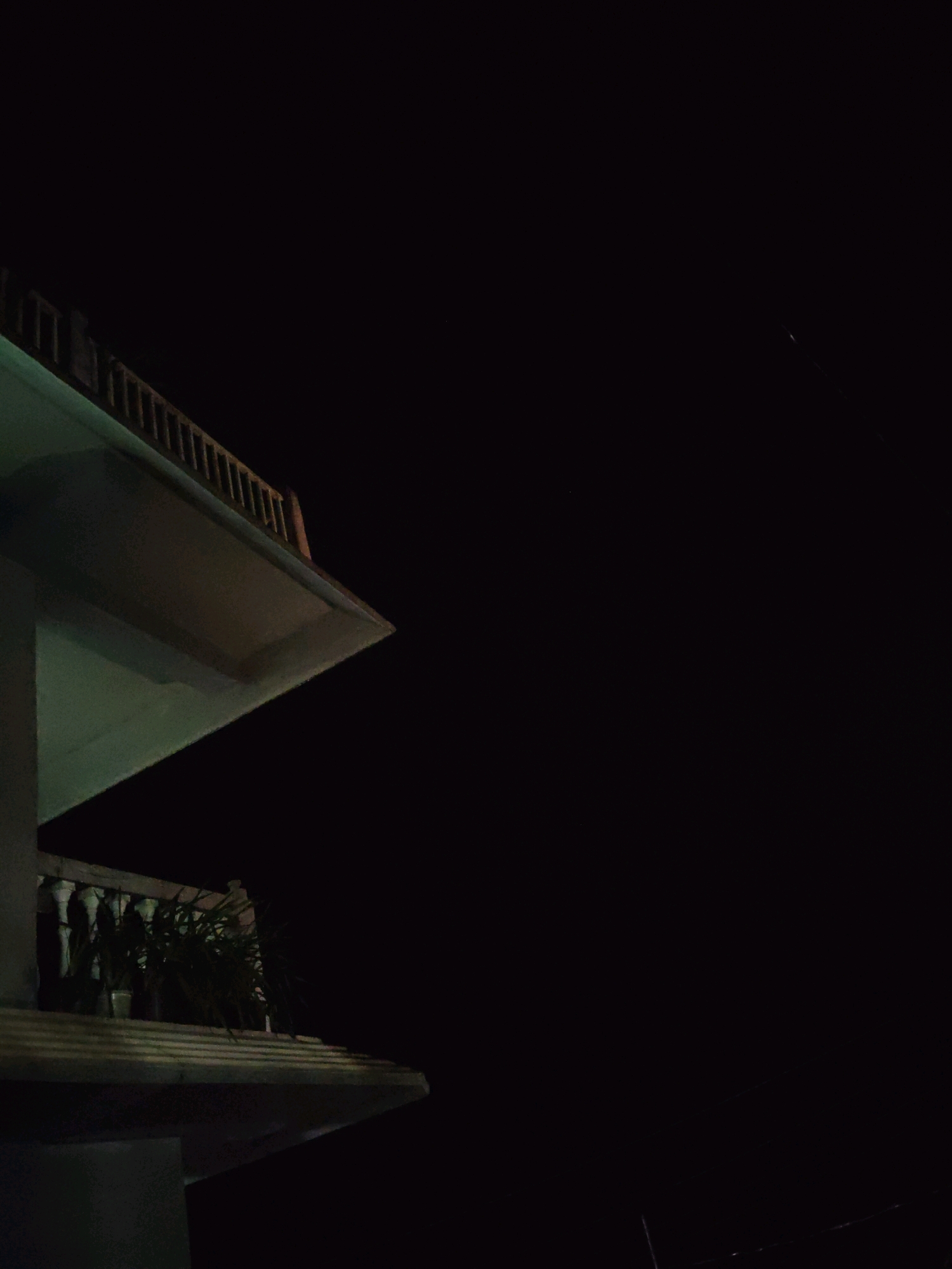 A low-angle, night-time photograph captures a portion of a modern building's exterior, characterized by its geometric design and balconies. The building, rendered in shades of grey and off-white, extends from the left edge of the frame, with its stark, clean lines contrasted against the deep black expanse of the night sky.
The upper balcony, a prominent architectural feature, juts out from the building. It is bordered by a light-colored railing composed of numerous vertical spindles and a darker horizontal top rail. The underside of this balcony is illuminated by a faint, cool green light, suggesting an artificial light source.
Below the upper balcony, another balcony is visible, also featuring a similar railing design with white, rounded balusters and a darker top rail. This lower balcony is adorned with several potted plants, their dark green foliage contrasting with the lighter structures around them. The plants appear to be a mix of leafy and grass-like species.
The background is dominated by the complete darkness of the night sky. A single, faint streak of light, possibly a distant airplane or satellite, is barely discernible in the upper right quadrant of the image. The overall atmosphere is one of quiet stillness, with the artificial lighting of the building providing the only illumination against the nocturnal darkness.