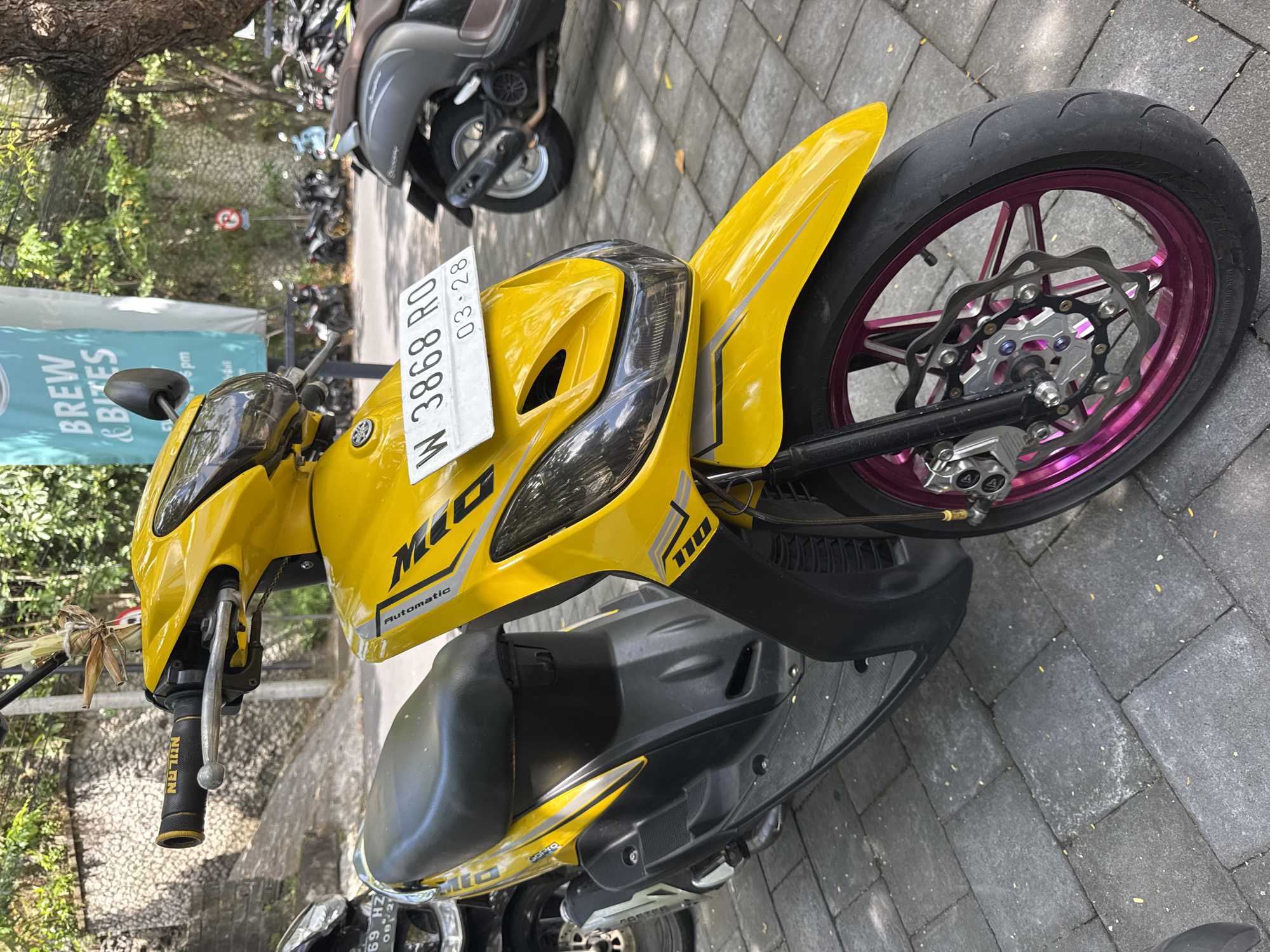 The image shows a yellow Yamaha Mio scooter parked on a brick paved surface. The scooter has a license plate that reads "W 3868 RO" with an expiration date of March 2028. The front wheel features a purple rim and a silver brake disc. A sign with "BREW & BITES" is visible in the background, along with other parked scooters. The photo was likely taken in Kuta Selatan, Indonesia, due to the vehicle registration and environment.