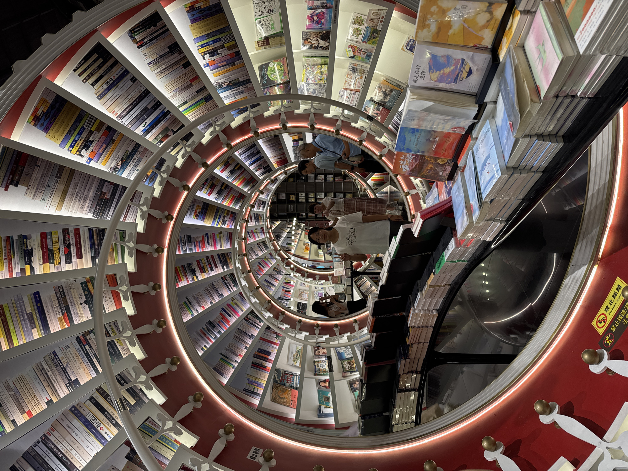This image is an upside-down, unusual perspective of a spiral bookshelf, likely taken within a bookstore in China’s Bao'an District. The shelves are stocked with numerous books, predominantly Chinese titles. Within the central opening of the spiral, reflections of people and the store's interior are visible. The structure features a red and white design, with a lighted rim illuminating the curving shelves. A sign warns about climbing or trampling. The image creates a visually confusing, almost surreal effect with the distorted orientation.