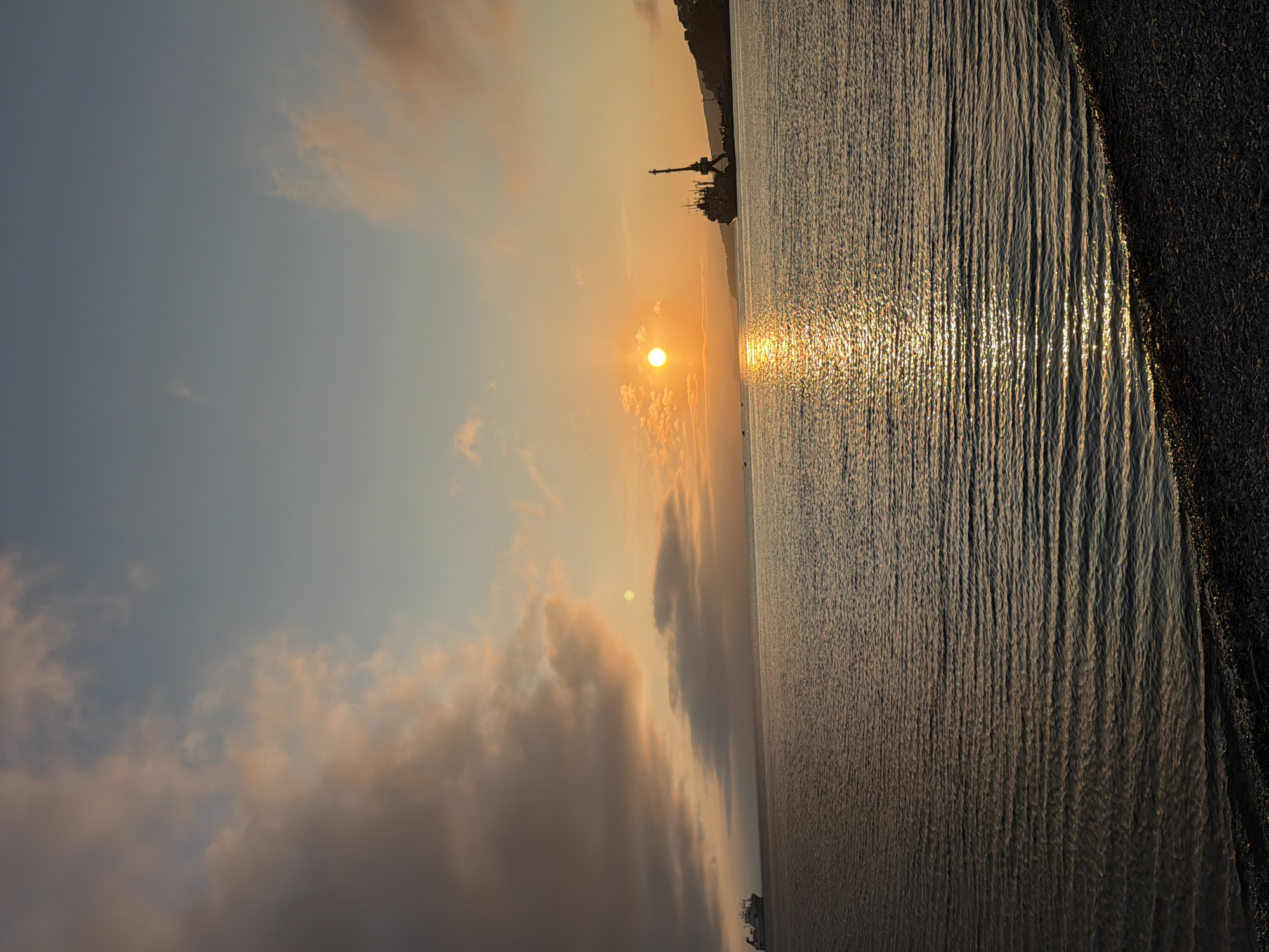 The image depicts a serene coastal scene at sunset in Petropavlovsk-Kamchatsky, Russia. The sky transitions from a soft blue at the top to warm oranges and pinks near the horizon, illuminated by a bright, setting sun. Wispy clouds, tinged with the sunset's glow, dot the sky. The calm sea mirrors the sky's colors, with gentle ripples creating a shimmering effect from the sun's reflection. In the distance, a large cargo ship is visible on the left, while on the right, a port area is discernible with a prominent crane and other vessels docked along the shoreline, which is bordered by a rocky beach in the foreground. The overall atmosphere is peaceful and tranquil.