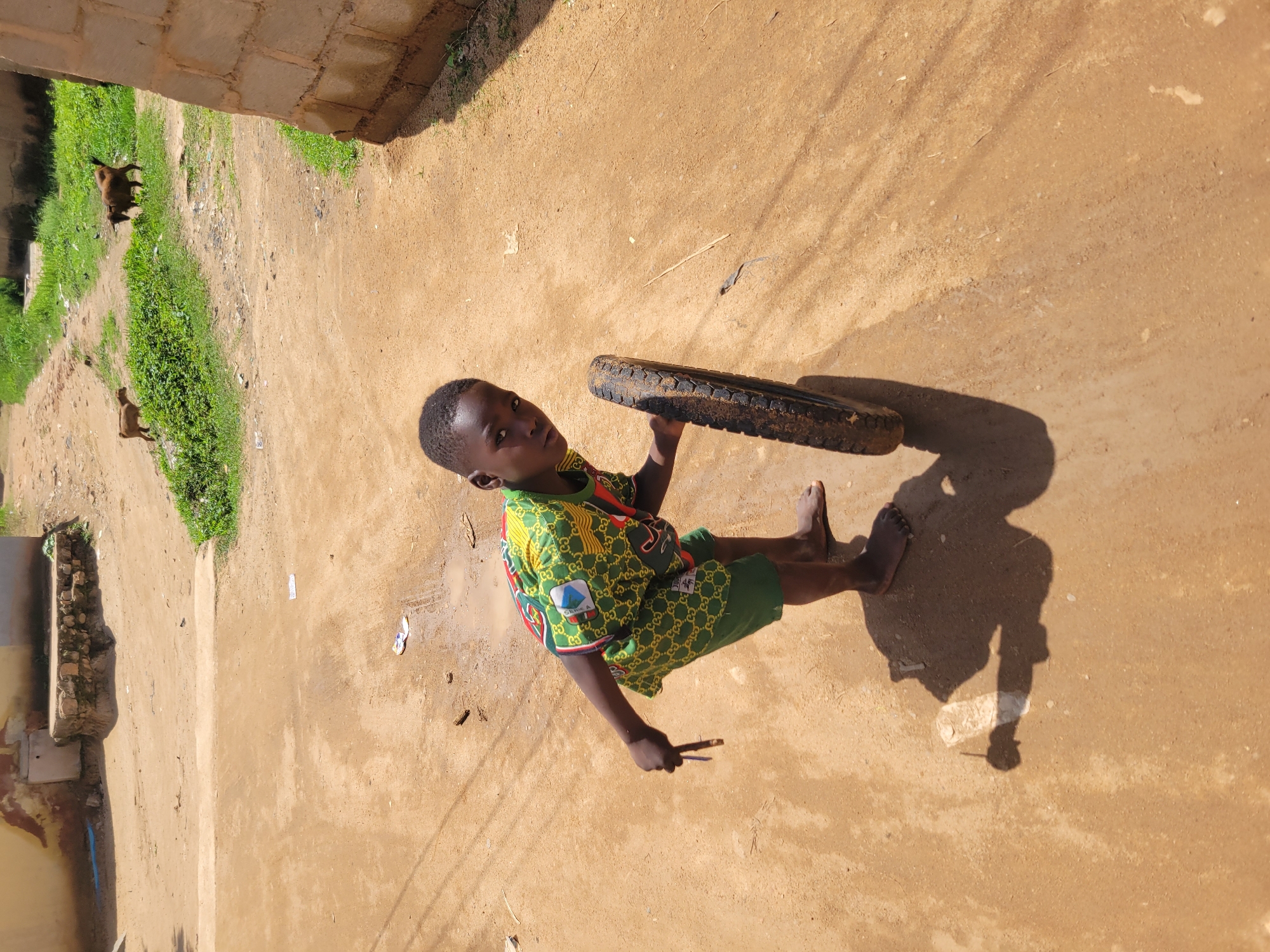 A young boy stands on a dirt path in Bukuru, Nigeria, holding a tire and a small stick. He wears a green and yellow patterned shirt and green shorts, with his right arm bent and holding the stick, while his left hand rests on the tire. The tire is black and worn. The ground is a mix of dry dirt and patches of mud, with some scattered debris including small pieces of paper and twigs. In the background, two goats are visible on a grassy area. The environment appears to be an outdoor, semi-urban setting with a building constructed of concrete blocks to the right of the frame. The lighting suggests it is daytime, with strong shadows cast on the ground.