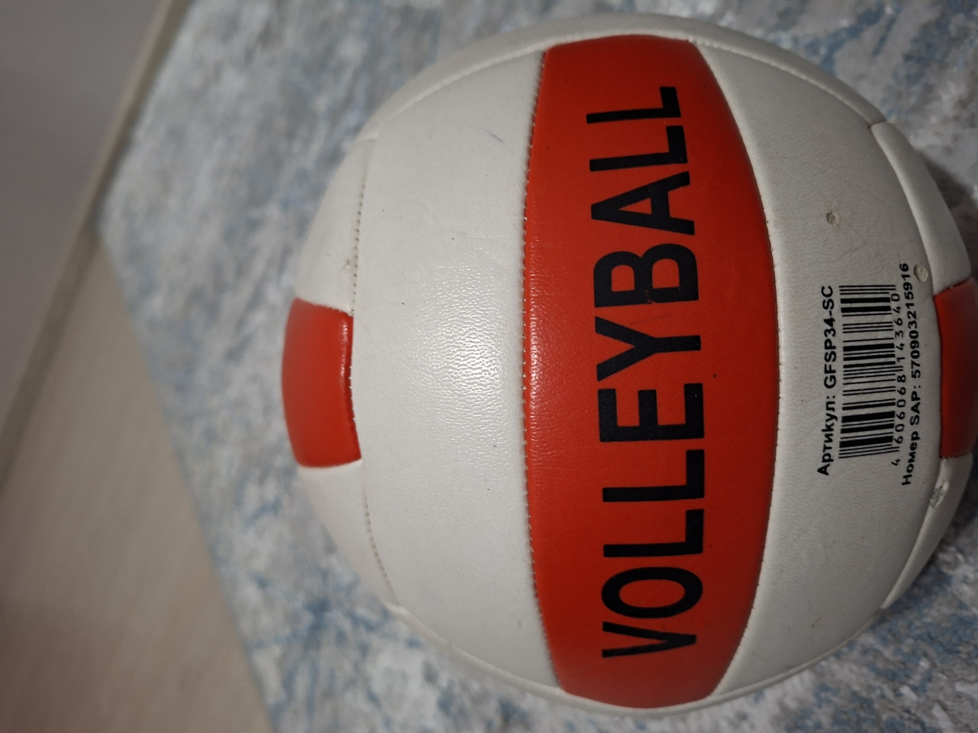A close-up photograph shows a white and red volleyball sitting on a light blue and gray patterned surface, likely a rug or carpet. The word "VOLLEYBALL" is printed in large, black, sans-serif font across a red stripe on the ball. To the right of the red stripe is a barcode with accompanying text in what appears to be Russian, including "Артикул: GFSP34-SC" and "Номер SAP: 570903215916". The background is slightly out of focus.