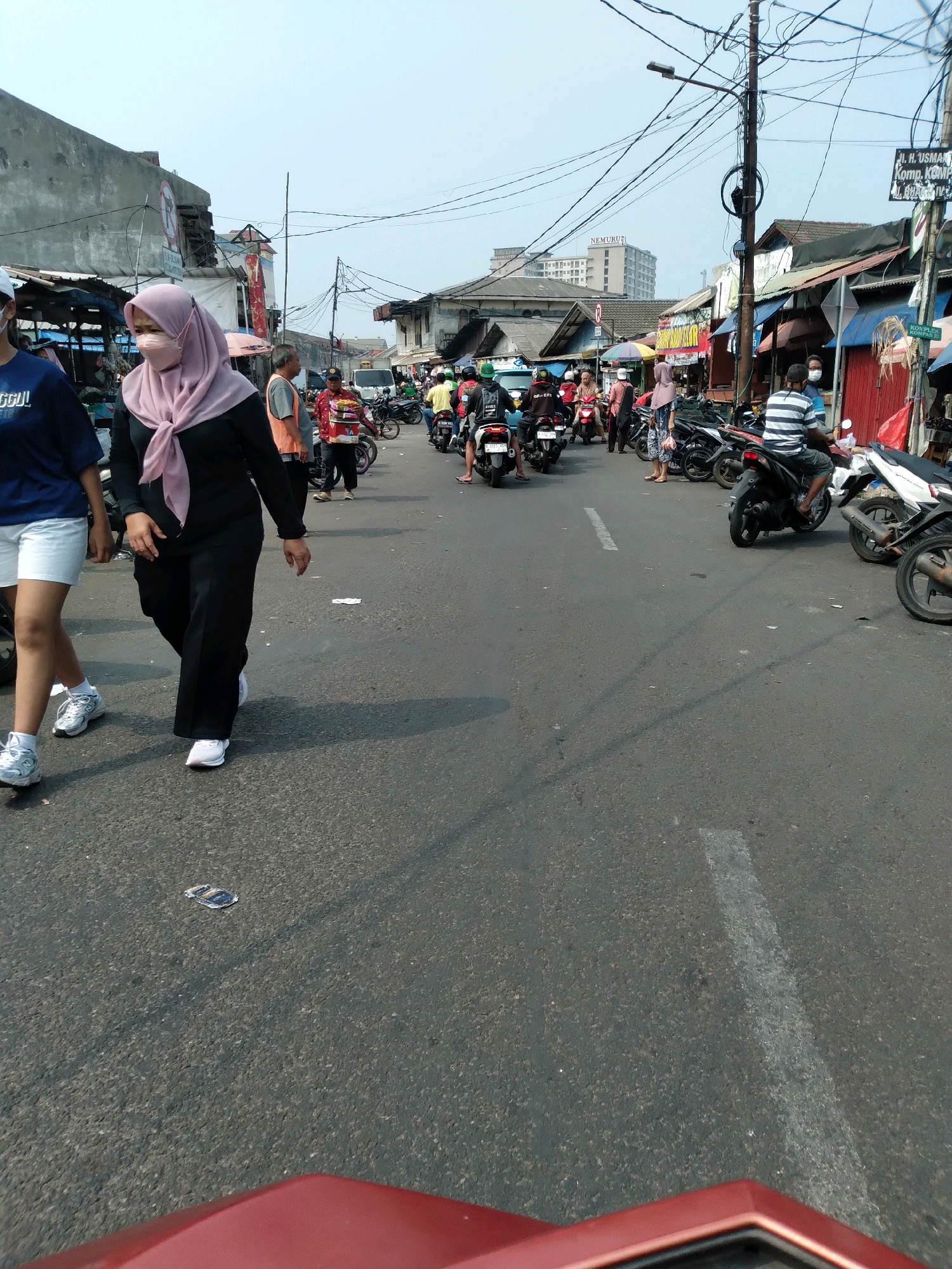A street scene in Tangerang, Indonesia, captures a busy marketplace environment. A woman in a pink hijab and black attire, wearing a face mask, walks across the asphalt road, her white sneakers visible. To her left, another person in a blue shirt and white shorts also walks on the road, wearing white sneakers. The road is lined with parked motorcycles and stalls with awnings, typical of a street market. Several people are seen on motorcycles, some wearing masks. In the background, buildings, power lines, and a taller building in the distance are visible under a bright sky. The ground is littered with a few pieces of trash.