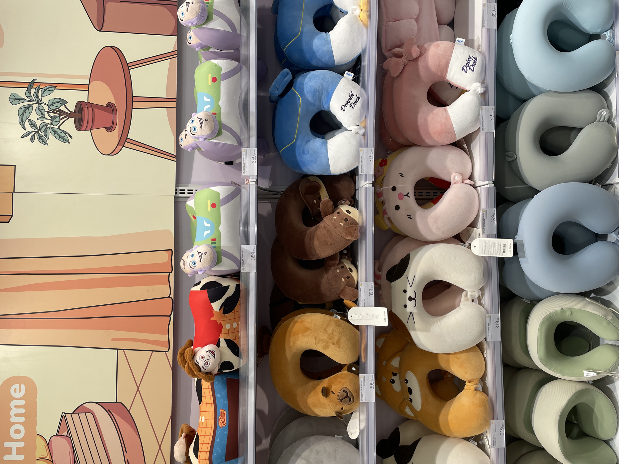 The image displays shelves in a retail store, likely a department store or a specialty shop, filled with various travel pillows. The store appears to be located in Davao City, Philippines, as indicated by the prices shown in the image, which are in Philippine Pesos (PHP).
The top shelf features novelty travel pillows designed to resemble characters from the Toy Story franchise, including Woody and Buzz Lightyear. These are positioned in front of a wall graphic depicting a home interior with a plant on a table.
The second shelf showcases a collection of animal-themed travel pillows, primarily in U-shapes. There are pillows designed to look like bears in brown and tan colors, and a blue pillow with "Donald Duck" embroidered on it. These pillows have price tags attached.
The third shelf continues the theme of animal-shaped travel pillows, with designs resembling a Shiba Inu, a cat, and a rabbit, all in soft, pastel colors.
The bottom shelf displays solid-colored U-shaped travel pillows in various shades of green, gray, and blue.
The overall environment is brightly lit and organized, typical of a retail display designed to attract customers. There are no people visible in the image, and the focus is entirely on the merchandise.