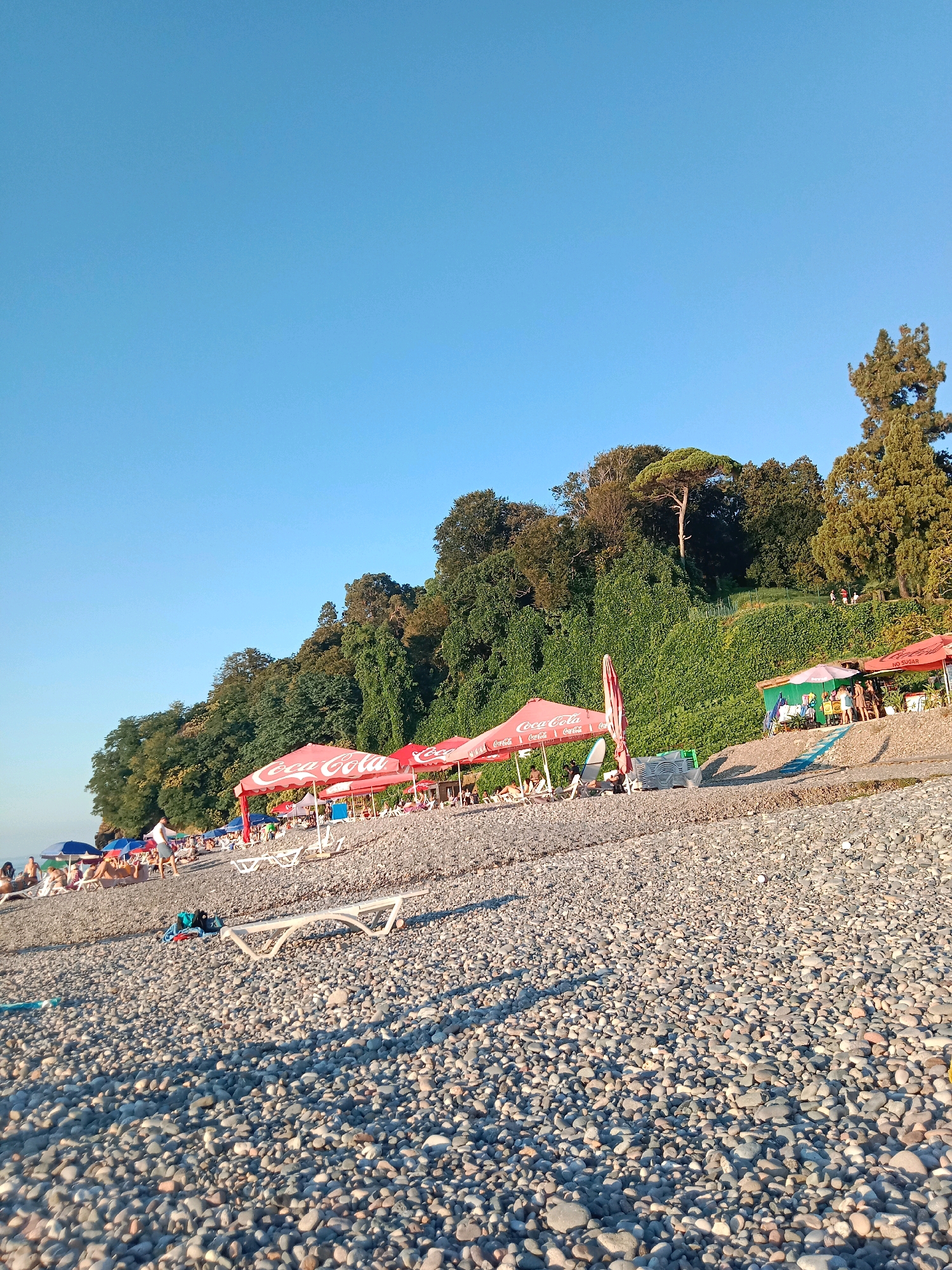 A wide-angle, eye-level shot captures a lively pebble beach in Batumi, Georgia, under a clear blue sky. The foreground is dominated by smooth, grey, and brown pebbles that fill the lower half of the frame. Scattered amongst the pebbles are several white plastic lounge chairs, some in use by beachgoers, and a few black and blue bags.
In the mid-ground, a line of red and white beach umbrellas with "Coca-Cola" branding provides shade for patrons. Behind these umbrellas, a vibrant green hillside rises, densely covered with lush trees and vegetation, creating a striking contrast with the beach. A few people are visible sitting or standing under the umbrellas, enjoying the seaside atmosphere. Some individuals are also seen walking along the shore or relaxing on lounge chairs.
Further back, the hillside extends upward, with more trees and patches of green. A few small figures can be seen walking along a path on the hillside. The far right of the image features a small green structure, possibly a kiosk or changing room, with a few umbrellas and people gathered around it. The overall scene depicts a popular tourist spot on a sunny day, with the natural beauty of the Georgia coastline forming a scenic backdrop.