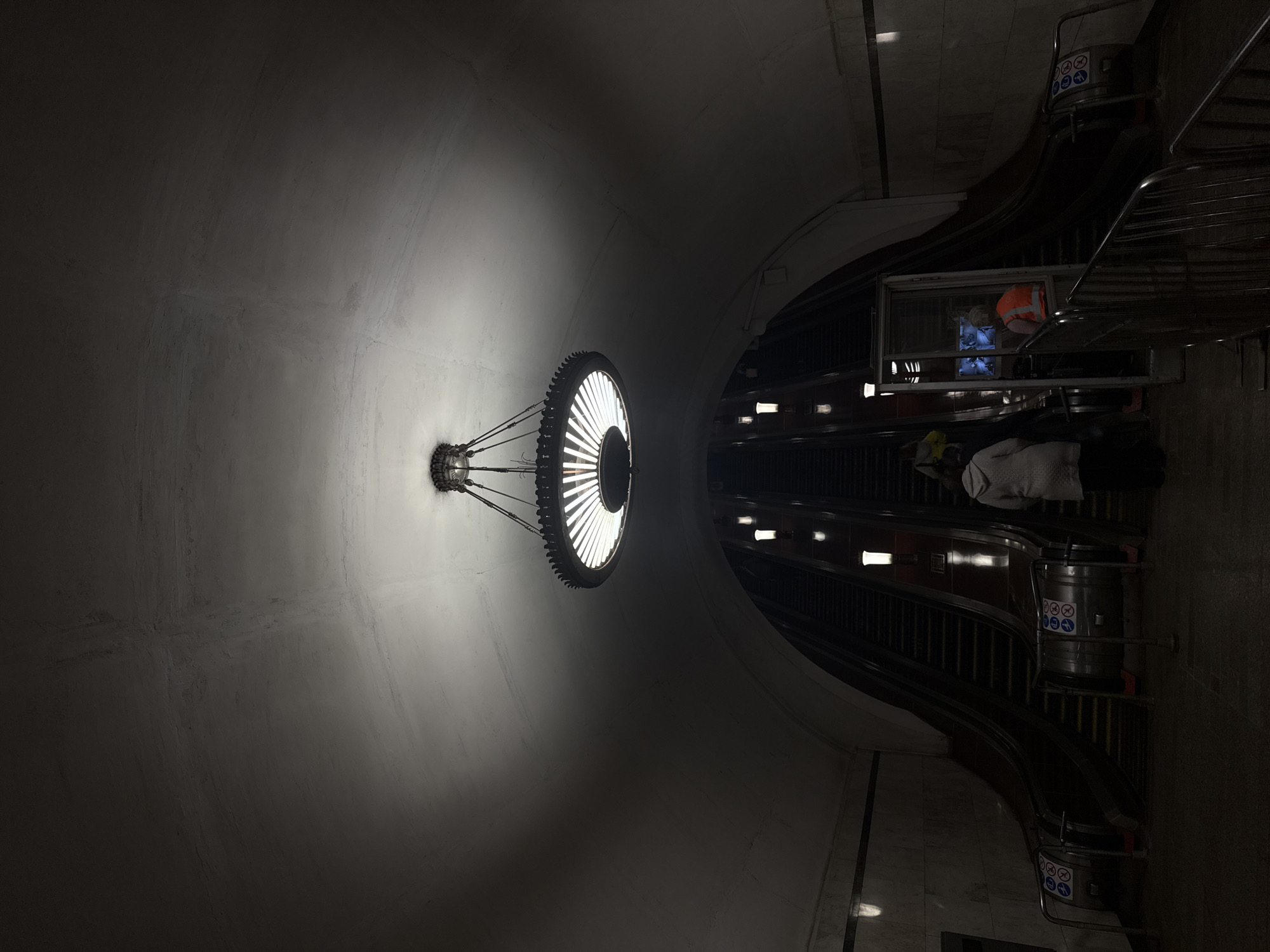 A dimly lit interior of the Kiyevskaya Metro station in Moscow, Russia, features a large, circular light fixture suspended from the curved ceiling. An escalator rises into the station from below with at least two people on it. One worker wearing a safety vest is in the escalator's service area. A safety sign is affixed to the end of the escalator.