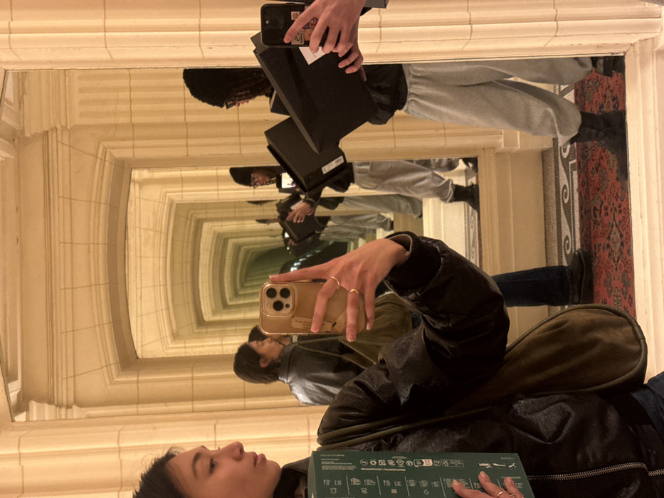 The image displays a selfie taken in a mirrored hallway, creating an infinite reflection effect within the Palais Galliera in Paris, France. In the immediate foreground, a person with dark hair, wearing a dark jacket and an olive-green shoulder bag, holds a gold-colored smartphone in their right hand, angled to capture the scene. Rings are visible on their right hand's fingers. Their left hand holds a green rectangular object. Reflected behind this individual is another person, partially obscured, wearing a dark jacket. Further down the mirrored hallway, a third individual is repeatedly reflected, diminishing in size. This person wears a dark beanie, glasses, a dark hoodie, light grey sweatpants, and dark boots, and holds two black rectangular objects. The architectural environment features light-colored walls with classical pilasters, arched openings, and a patterned floor, consistent with the museum's interior design.