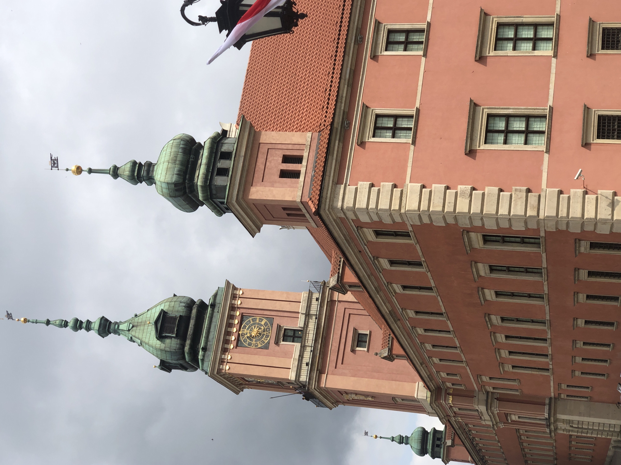 The image shows the Royal Castle in Warsaw, Poland, under a cloudy sky. The castle is a large, pinkish-orange building with a red tiled roof. It features two prominent green-domed towers with spires, one of which has a large clock face. The architecture is classic European, with ornate windows and decorative stonework. A black lantern with red and white flags hangs in the upper right corner of the frame. There are no people visible in the image. The overall impression is one of historical grandeur and imposing architecture.