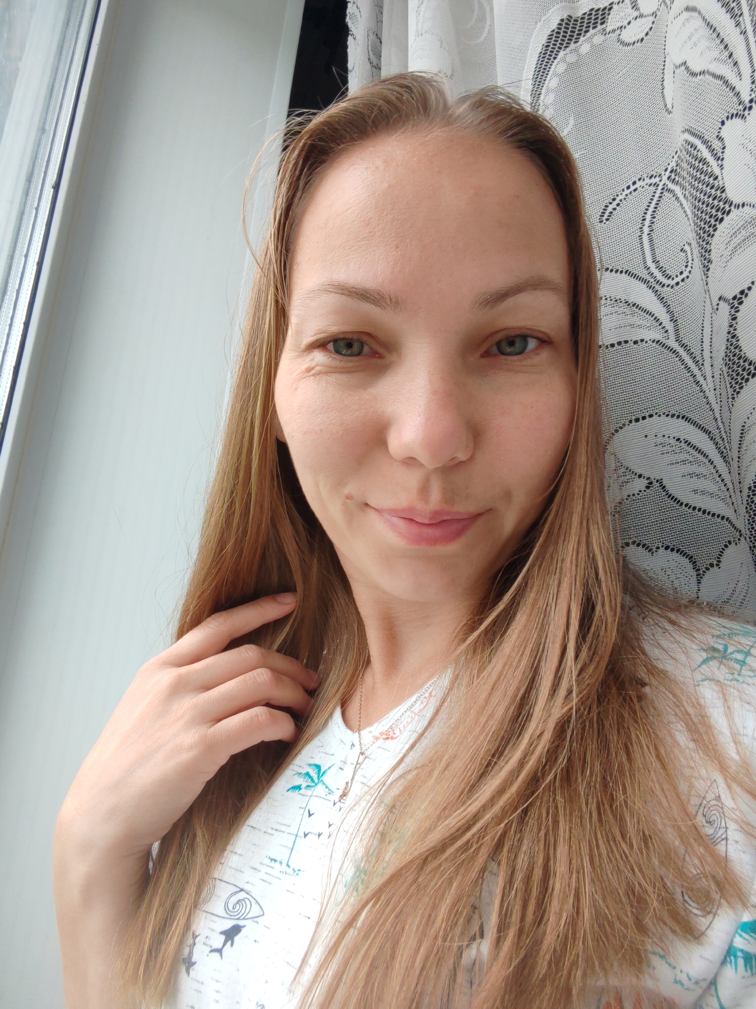 A medium shot photograph shows a Caucasian woman with long, light brown hair and green eyes. She is smiling faintly and looking towards the camera, with her left hand touching her hair. She is wearing a white t-shirt with a tropical print featuring palm trees, dolphins, and waves. Behind her, a white lace curtain is visible, and to her left, a window frame with a white sill. The lighting suggests an indoor setting during the daytime, possibly near a window. No specific location in the city of Чухлэм, Russia, can be identified from the image.