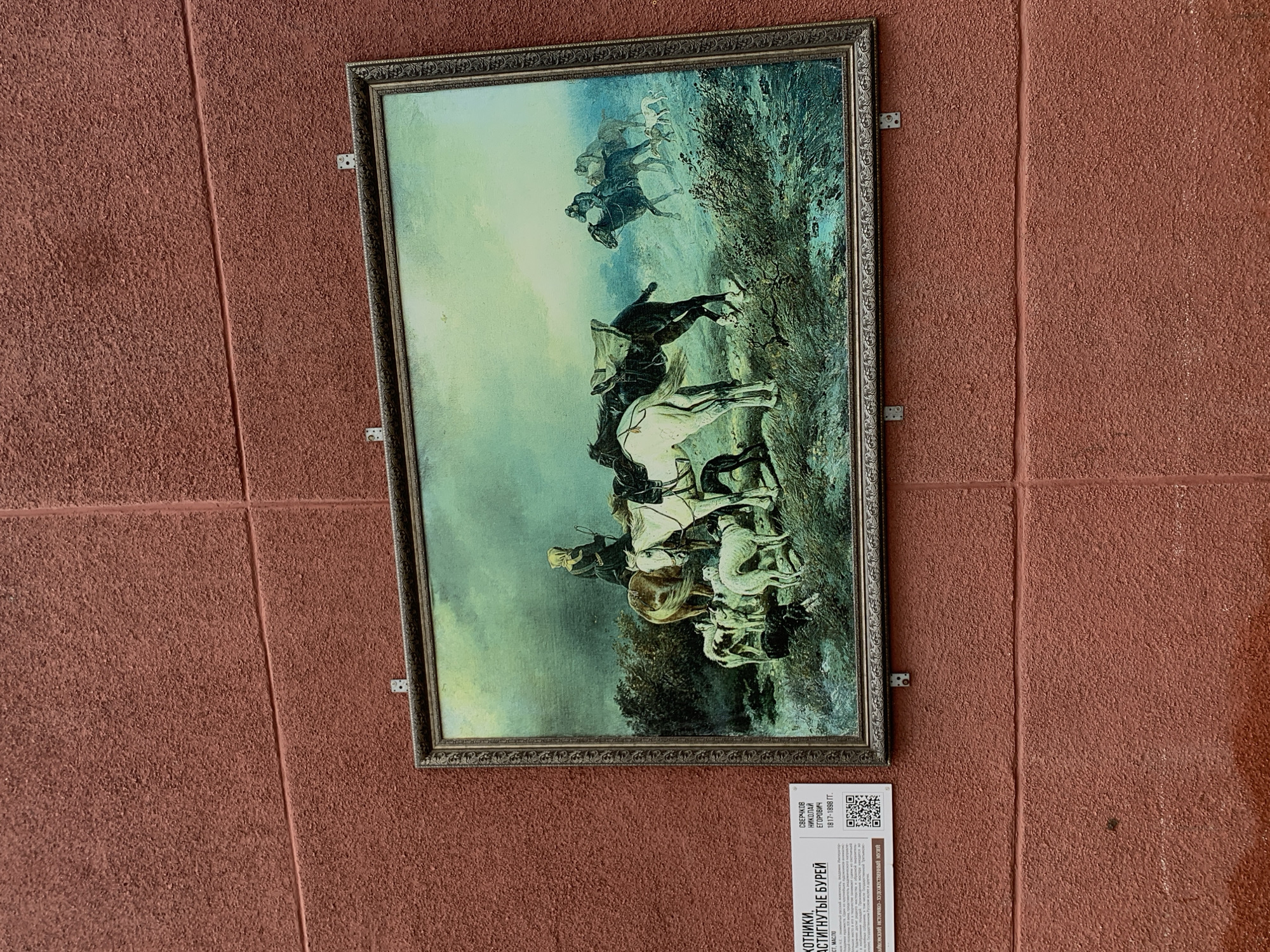 A framed painting hangs on a reddish-brown textured wall. The painting depicts a group of horses and riders in a field. The painting is titled "Huntsmen overtaken by the Storm" by Nikolai Egorovich Sverchkov. A white card is affixed below the frame, containing text in Russian, including the artist's name and the Baikovsky Historical and Art Museum. The painting is located in the city of Chaikovsky, Russia.