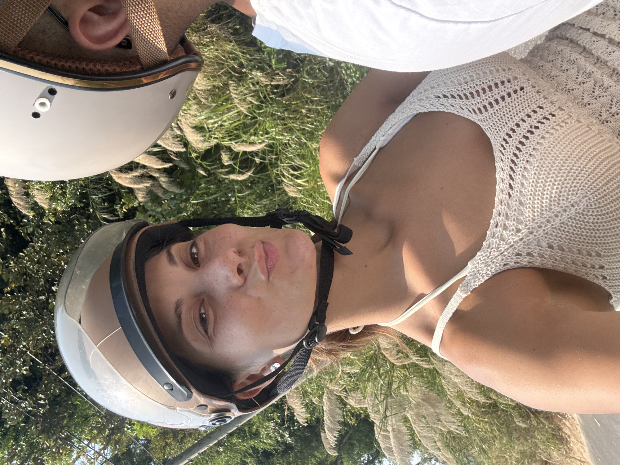 A selfie of two people on a scooter in Đà Nẵng, Vietnam. A woman with brown hair and a helmet with a transparent visor is facing the camera. She has a white top. Beside her, a man with a helmet is visible only partially. The background shows greenery and tall grasses.