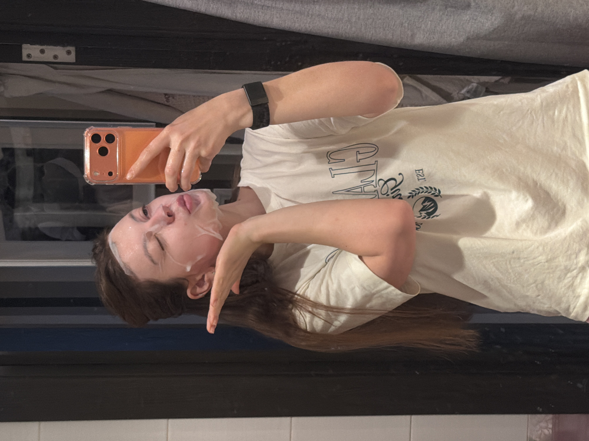A young person, likely female, is captured in a mirror selfie indoors in Khimki, Russia. They are wearing a white or off-white t-shirt with dark, reversed text that appears to read "CITY" and "EST. 2" followed by a logo featuring a mountain and leaves. Their face is covered with a white, foamy or creamy face mask, indicating a skincare routine. On their left wrist, a black fitness tracker or smartwatch is visible. The person holds an orange smartphone in a clear case with their right hand, using it to take the picture.
The subject's expression is playful or silly, with their eyes closed or squinting, and their tongue slightly extended. Their left hand (from the viewer's perspective) is posed with fingers pointing downwards. Long, dark brown hair cascades down the subject's back. The background, visible in the mirror's reflection, includes dark furniture or a door frame, hints of fabric (possibly a curtain or bedding), and white tiles at the very bottom, suggesting a domestic setting like a bathroom or bedroom. The image conveys a lighthearted moment of self-care.