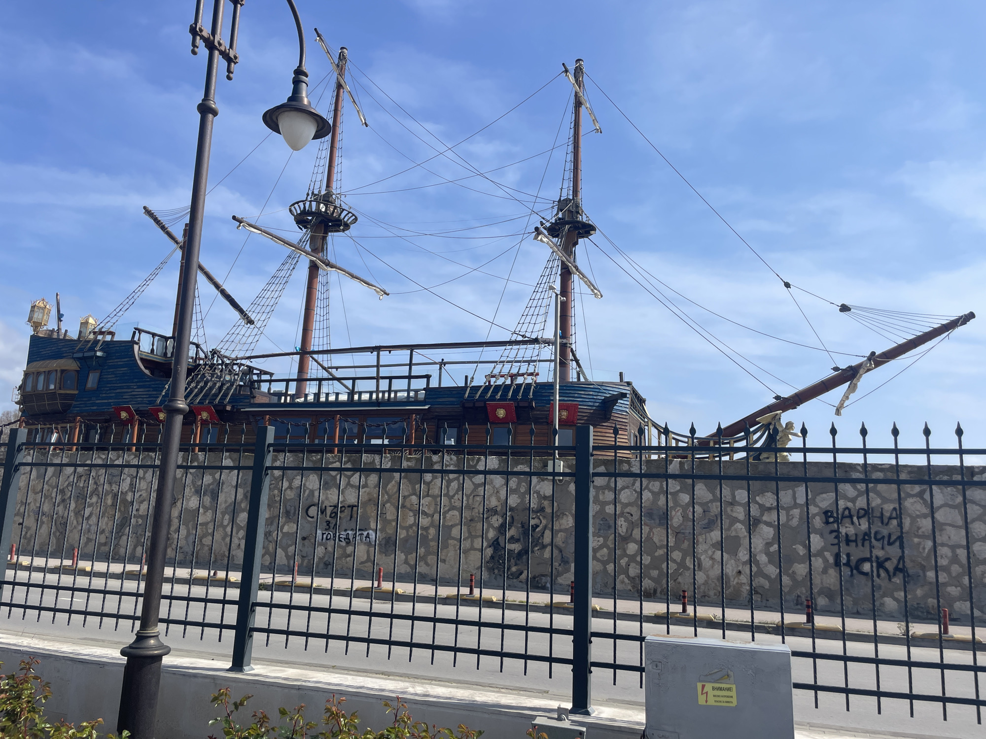 The image captures a scene in Varna, Bulgaria, featuring a large, static ship situated behind a metal fence and a low stone wall. The ship, reminiscent of a historical sailing vessel, has visible masts, rigging, and some deck structures. The hull appears to be constructed of wood and painted in various colors.
A street light is placed in the foreground to the left. The fence in the foreground is black, with vertical bars, and runs along the length of the image. Behind the fence is a stone wall, which serves as a backdrop for the ship. Graffiti is visible on the stone wall behind the fence.
The sky is blue with scattered clouds, suggesting a sunny day. The overall setting appears to be a public area, possibly a waterfront or tourist location in Varna, given the presence of the decorative ship and the public infrastructure. A utility box is placed on the ground.