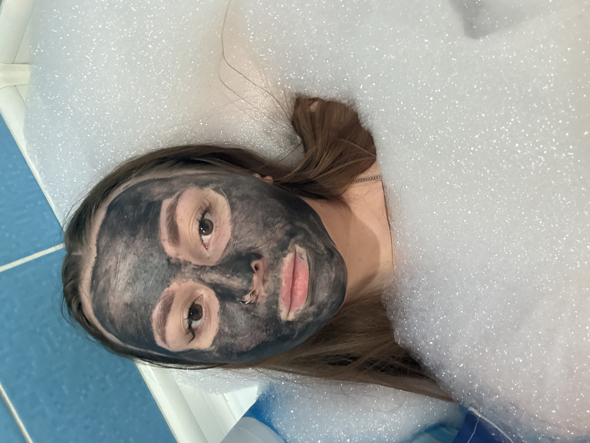 A young woman is depicted lying in what appears to be a bathtub filled with abundant white foam, likely a bubble bath, in Izhevsk, Russia. She is wearing a dark grey or black face mask that covers most of her face, leaving her eyes, nostrils, and lips exposed. Her brown hair is spread out around her head and partially submerged in the bubbles. She has a silver or metallic nose ring in her left nostril and a delicate chain necklace is visible around her neck. Her eyes are open and gaze directly upwards towards the viewer, appearing calm. The pinkish hue of her lips contrasts with the dark mask.
The setting suggests a bathroom, with a section of blue-tiled floor or wall visible in the lower left corner and a white, possibly ceramic, edge of a tub or surface around her head and upper body. The bubbles appear thick and glittery, catching reflections. The overall scene conveys an activity of self-care and relaxation, such as a spa treatment or bath time. No text is visible in the image.