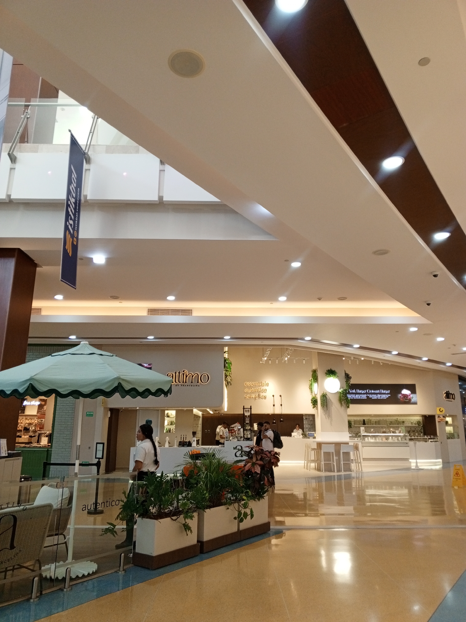 The image depicts the interior of a mall in Porlamar, Venezuela. Dominating the foreground is an outdoor seating area for a cafe named "Autentico", with a light green umbrella shading wicker chairs and a table. A female employee in a white shirt stands nearby. Behind her, the main shopping area is visible, featuring other food vendors such as "Attimo". Several shoppers are present. The environment is brightly lit with recessed lighting in the high ceiling. A banner labeled "Istikbal" hangs from the upper level. The floor is glossy and reflects the overhead lights. The overall scene suggests a modern, upscale shopping mall environment.