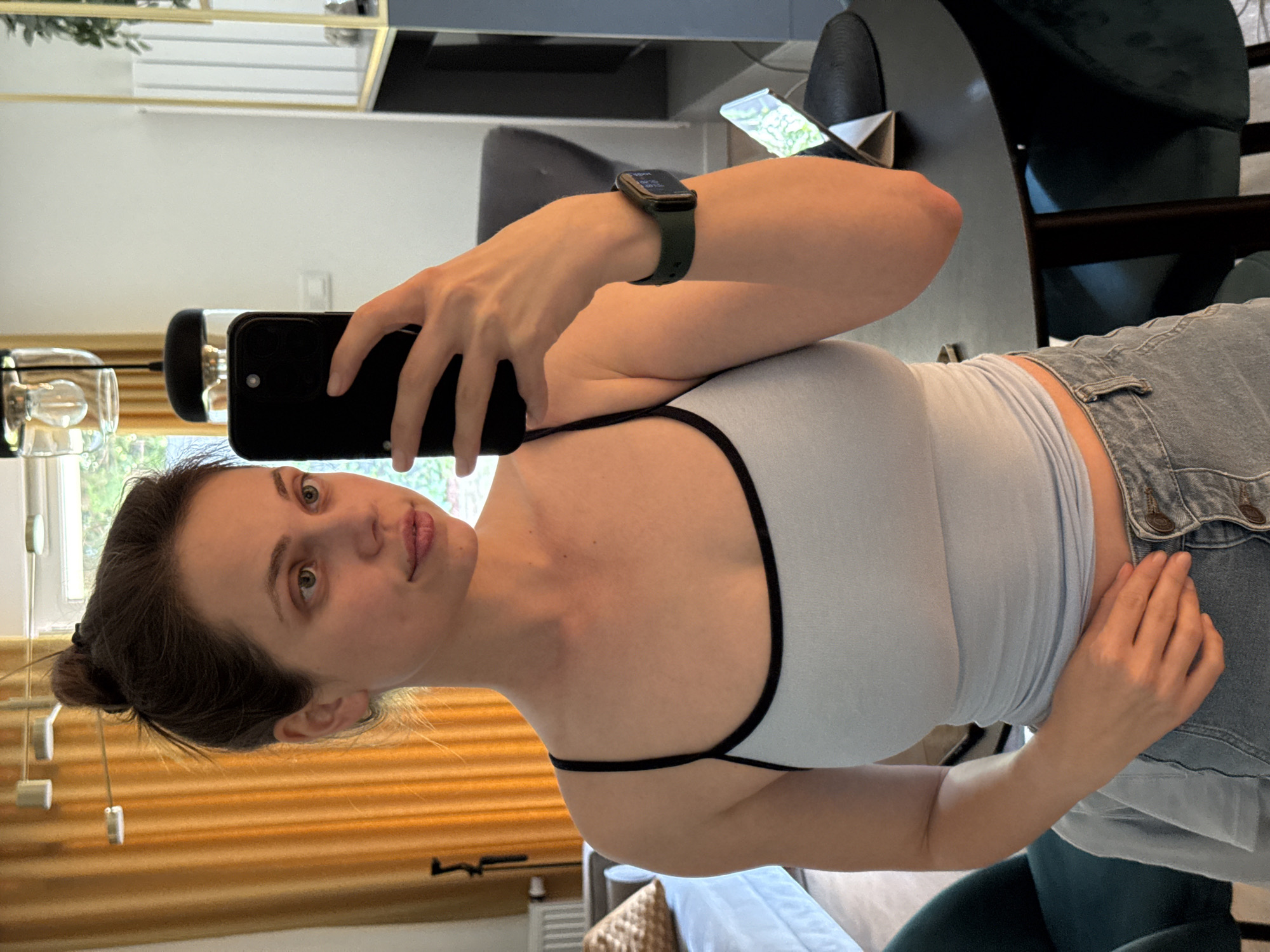 This media file features a young woman taking a mirror selfie. She is wearing a light blue tank top with black trim and light-wash denim shorts. A smartwatch with a dark green band is visible on her left wrist. Her hair is pulled back into a bun.
The setting appears to be indoors, possibly a home, with a blurred background that includes furniture and windows. The lighting suggests it could be daytime. The woman's expression is neutral as she looks into the camera. The image is taken from a slightly high angle, and the camera is held in her right hand. The overall mood is casual and candid. No visible text is present in the image.