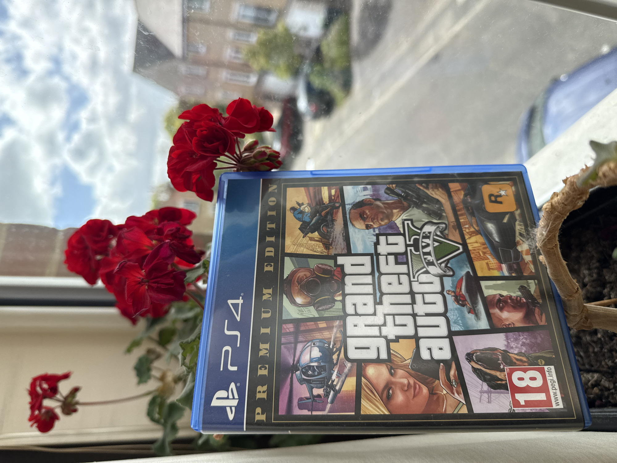 A medium-angle shot depicts a "Grand Theft Auto V Premium Edition" PS4 game case positioned on a windowsill next to a pot of red geraniums. The game case is partially obscured by the geraniums. Outside the window, buildings and vehicles are visible. The photo was taken in Great Yarmouth, United Kingdom.