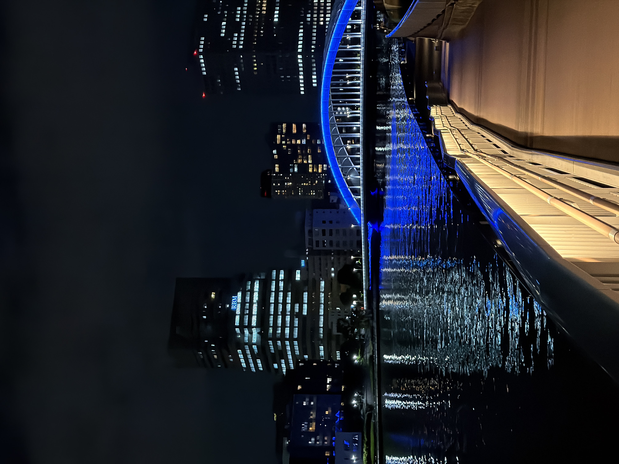 The image captures a nighttime scene in Koto, Japan, featuring a view of a brightly lit bridge over a body of water, with illuminated skyscrapers in the background. The bridge, arching gracefully, is outlined with vibrant blue lights that cast a strong reflection on the dark water below. On the right side of the frame, a pathway with a railing, also illuminated, leads away from the viewer. The water is calm, with the lights from the city and the bridge creating a shimmering, abstract pattern on its surface. The skyscrapers are tall, modern structures, their windows glowing with white and yellow light, indicating activity within the buildings. The sky is dark, suggesting it is late at night. There are no people visible in the frame. The overall impression is one of a serene and modern urban nightscape.