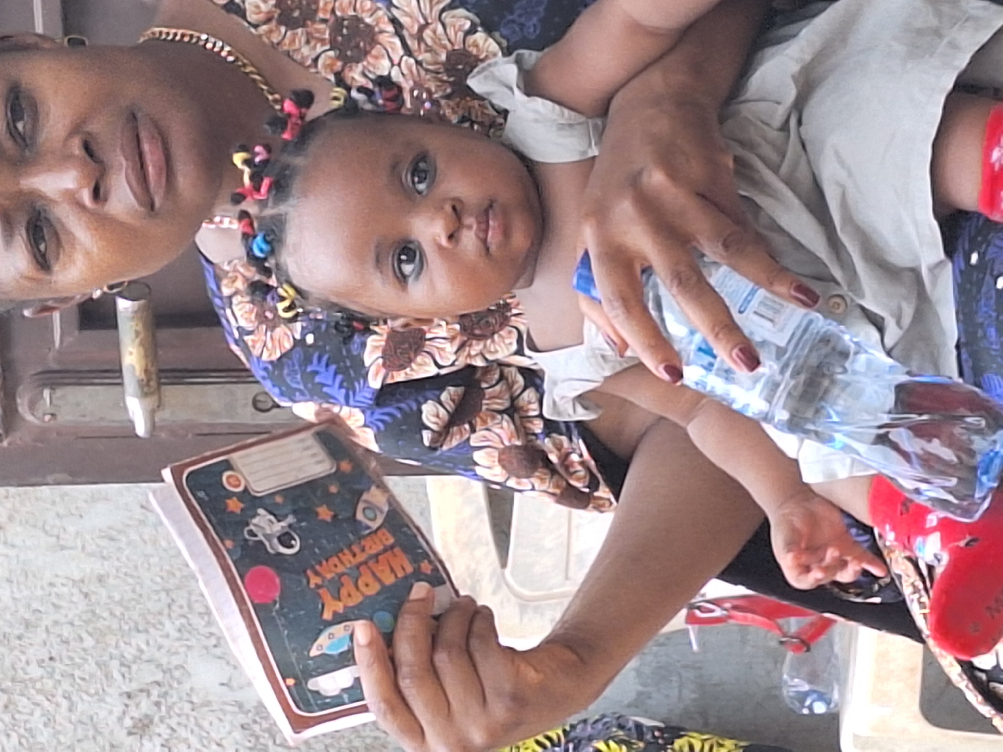 In an indoor setting in Sabon Tasha, Nigeria, a woman cradles a baby while seated. The woman has dark skin, wears a floral print top, and has a gold chain necklace. The baby is light-skinned, with braids adorned with colorful accessories. The baby wears a light-colored dress and red shoes. The woman holds a water bottle in one hand and a notebook with "Happy Birthday" printed on the cover in the other. The background includes a doorframe, and the floor is visible.