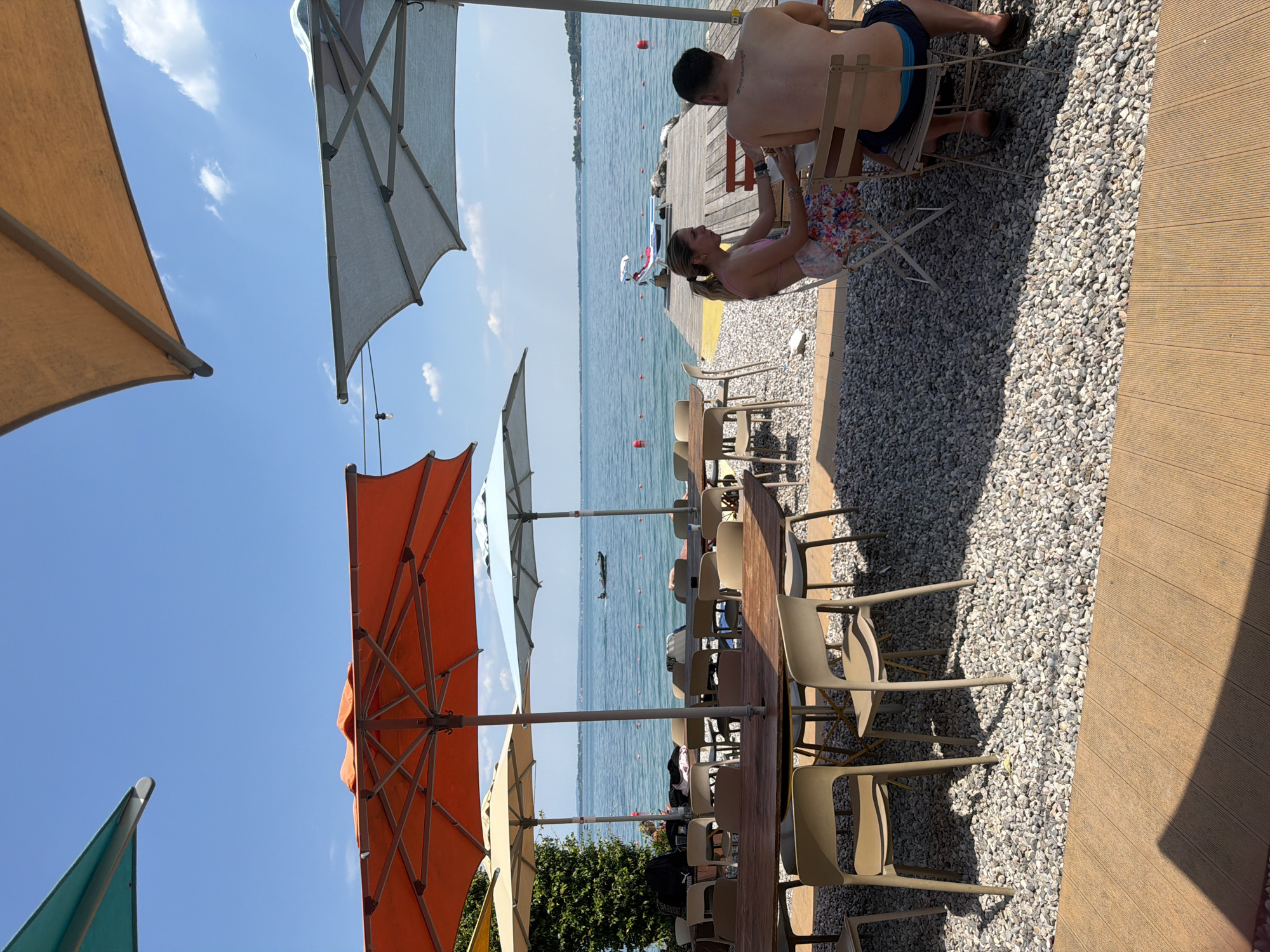 This is a daytime, eye-level, outdoor shot of a cafe situated on a lakeshore in Padenghe sul Garda, Italy. The scene is dominated by large, colorful umbrellas providing shade over tables and chairs. A couple is seated at a table, with a woman wearing a floral-patterned skirt and pink bikini top, facing a man with his back to the camera, wearing blue swim trunks. They appear to be engaged in conversation. In the background, a vast body of water, likely Lake Garda, stretches out under a clear blue sky. A wooden dock extends into the water, with a red and white lounge chair visible on it. A small boat with two people is present on the lake, and buoys mark the water's edge. The ground in the foreground is covered in small, light-colored stones, with a wooden deck in the bottom right corner. The overall atmosphere is relaxed and summery.