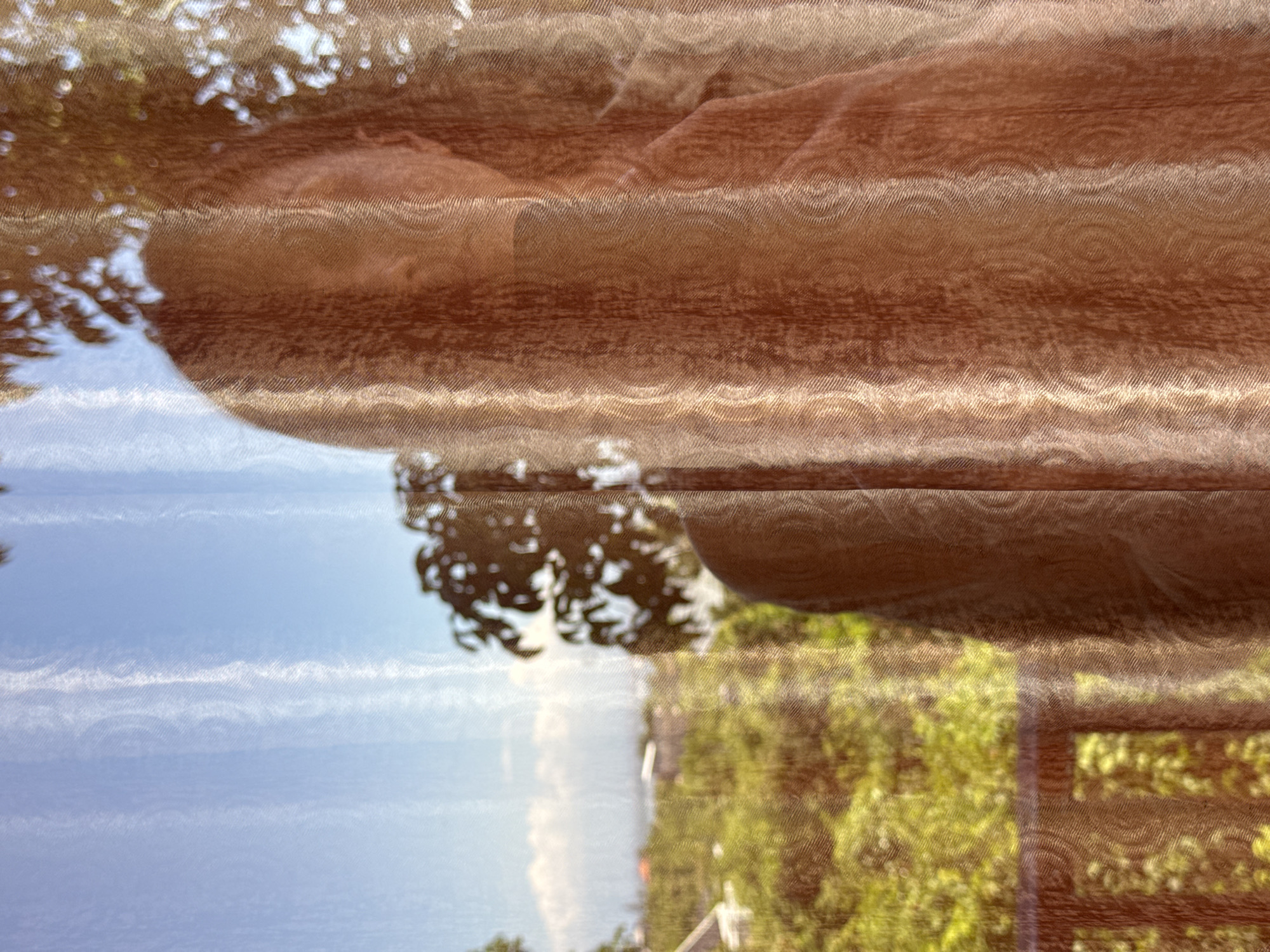 The image shows a scene viewed through a sheer curtain, likely indoors. The foreground is dominated by the textured, brown fabric of the curtain. Through the curtain, a view of the outdoors is visible.
The outdoor scene includes a blue sky with wispy white clouds, suggesting a bright day. Some trees with green foliage are also visible. The location is identified as Kuta Selatan, Indonesia. No people or interactions are directly discernible. The image primarily captures a static scene, emphasizing the effect of the curtain on the view.