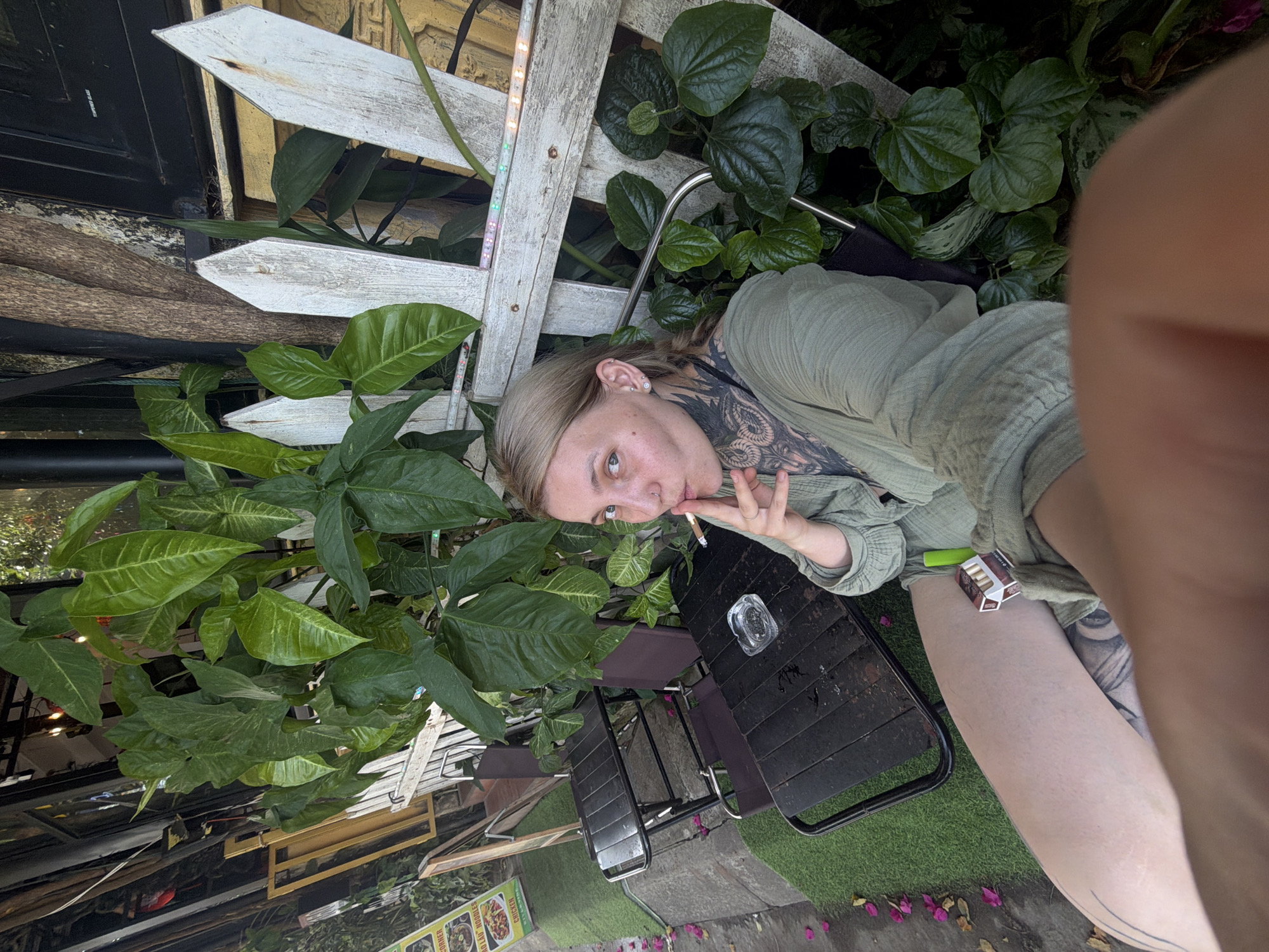 The image captures an individual taking a close-up selfie or being photographed in an outdoor or semi-outdoor setting. The subject, a light-skinned person with light-colored hair and blue/grey eyes, is positioned in the upper right, gazing directly at the camera. They wear a loose, light green long-sleeved garment and display prominent black ink tattoos on their neck/shoulder and upper left thigh. They are holding a lit cigarette to their lips, with a pack of L&M cigarettes and a light green disposable vape device resting on their thigh.
The setting is lush with various green plants, including large-leafed specimens and a vine growing on a weathered white wooden picket fence that forms the background. A string of small, partially lit LED lights is attached to the fence. In front of the subject are black metal tables and chairs, with a glass ashtray containing cigarette butts visible on one table. The ground is a mix of artificial green turf and pavement, with scattered pink flower petals. In the deeper background, a reflective surface shows hints of an interior or building structure.
The scene suggests a casual environment, likely a cafe or restaurant patio in Đà Nẵng, Vietnam, as indicated by the Vietnamese text on a partially visible menu sign in the lower left background. The sign advertises "Mì Xào Giòn," "HỦ TIẾU," and "NOODLES," with options for "CHICKEN," "BEEF," and "PORK." The lighting suggests it is daytime, possibly late afternoon. The individual's expression is direct and somewhat pensive.