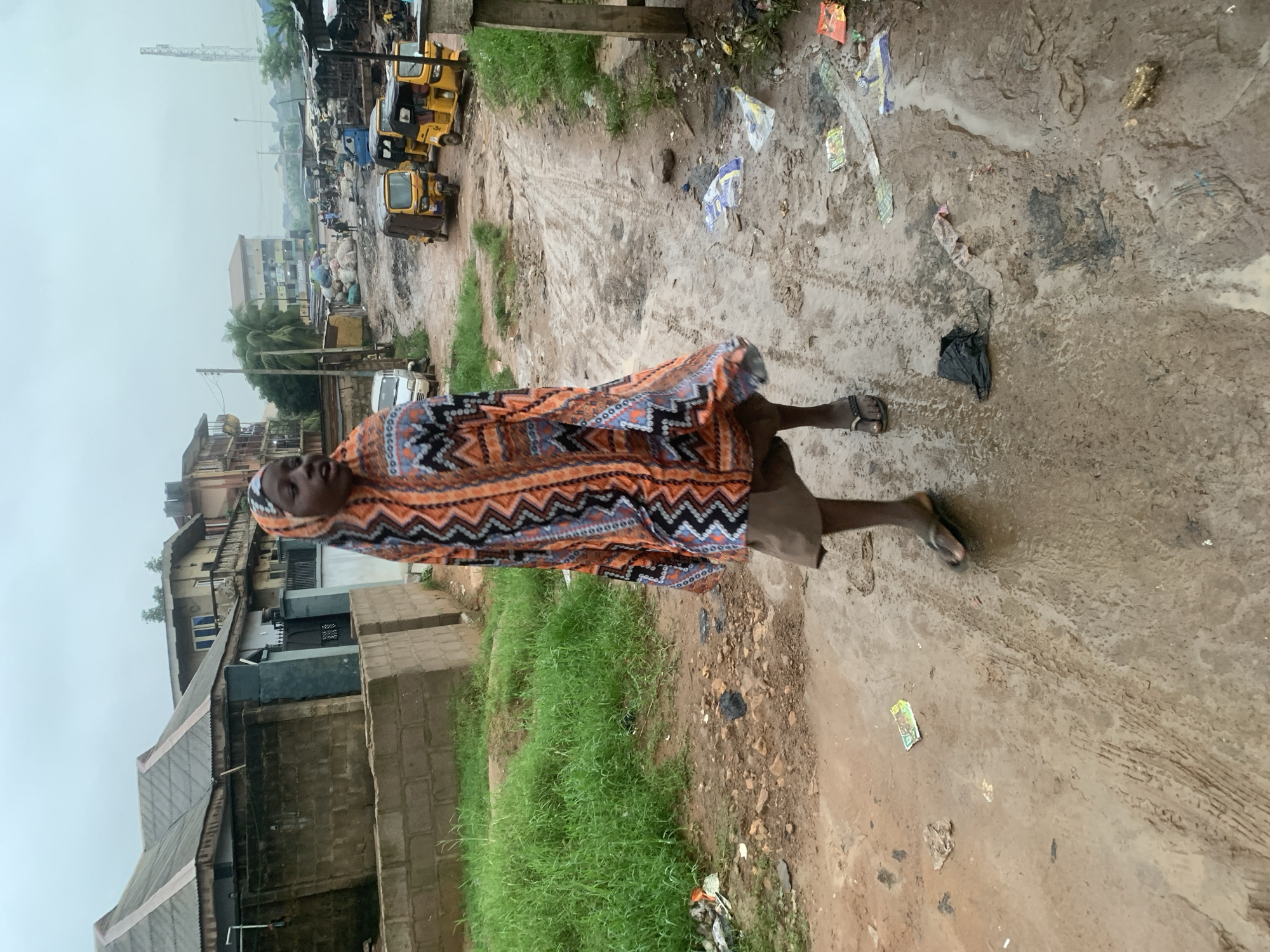 A young person wearing a colorful headscarf and patterned wrap walks on a muddy, unpaved road in Shagamu, Nigeria. The road is littered with debris, including plastic bags and papers. In the background, various buildings, including houses and market stalls, line the street. Several yellow auto-rickshaws, commonly known as "Keke Napep," are parked or driving on the road. The sky is overcast, suggesting a cloudy or rainy day. The scene depicts a typical street environment in a Nigerian city, likely a commercial or residential area.