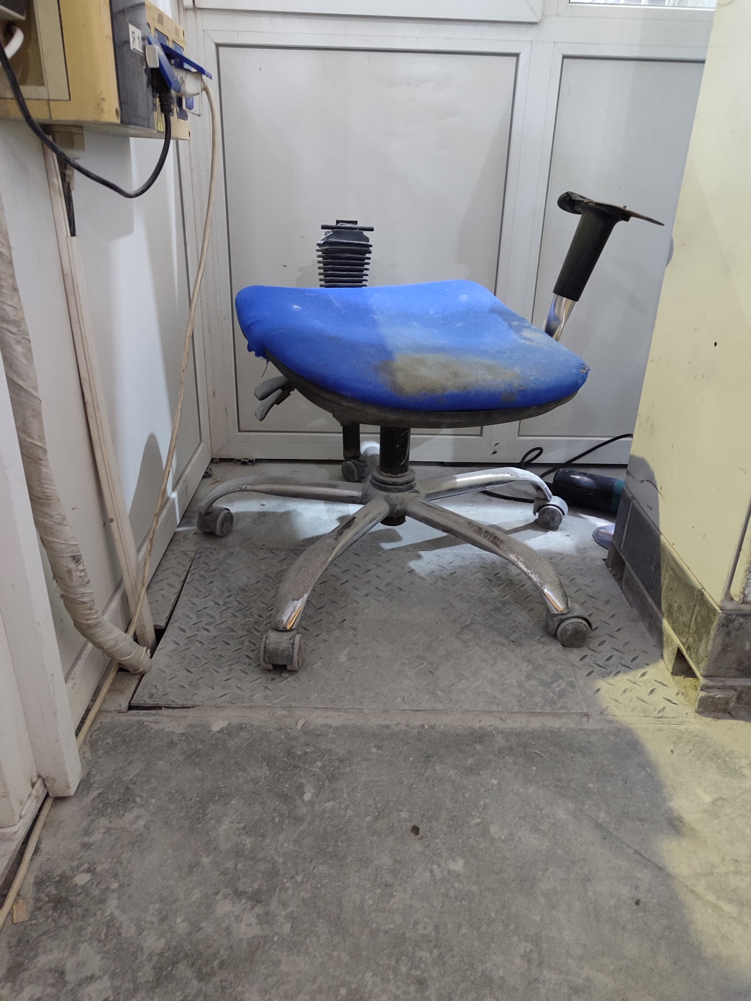 This image shows a blue, wheeled office chair in a utilitarian setting. The chair has a somewhat worn appearance, with visible dust and grime on its seat and metallic base. It is positioned on a diamond-plate metal surface, suggesting a workshop, industrial area, or perhaps a service space.
To the left of the chair, a yellow electrical box is mounted on a white wall, with several cords plugged into it. One cord is black and thicker, while another is thinner and off-white. The wall appears to be part of a partition or doorway. A white, plastic-wrapped pipe or conduit runs vertically along the left side of the image, obscuring some of the wall.
To the right of the chair, a portion of a light-colored, rectangular object is visible, possibly a piece of equipment or furniture. A black power cord from this object snakes across the floor.
The background consists of a white wall and a door or cabinet with vertical paneling. The lighting is somewhat harsh, casting shadows and highlighting the dusty surfaces. The overall impression is one of a functional, possibly under-maintenance, space. No people are visible in the image, and there are no obvious indicators of the time of day or weather conditions, beyond the artificial interior lighting. The location context of Dnipro, Ukraine, does not provide specific visual cues in this image. There is a label on the yellow electrical box with the number "9 1" visible.
