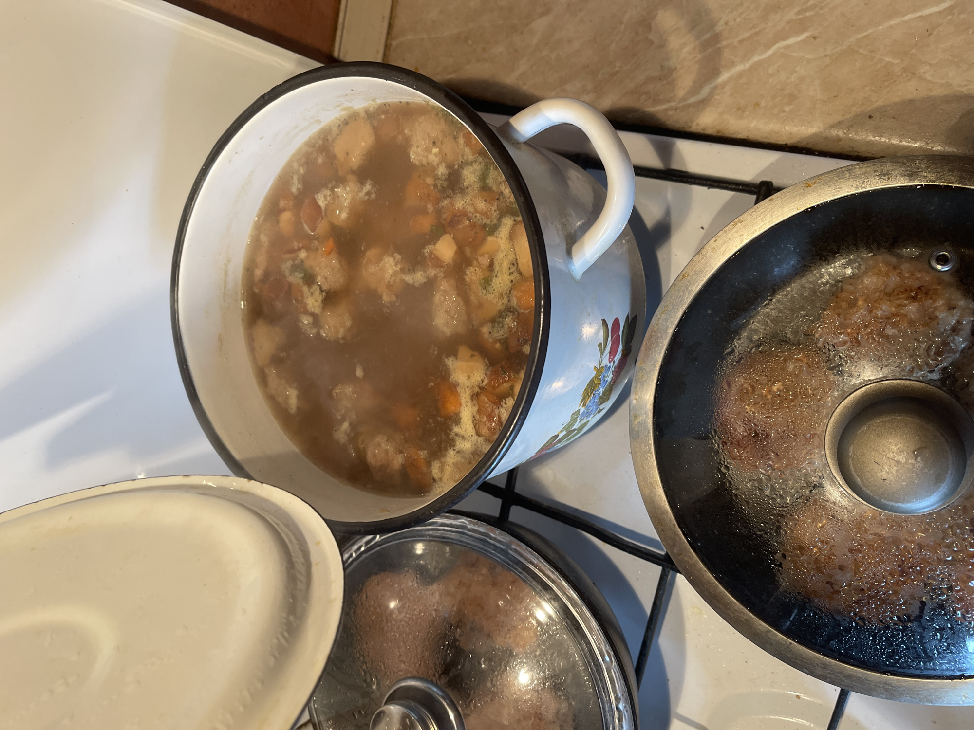 In an indoor domestic kitchen environment, possibly located in Shcherbinka, Russia, three cooking vessels are visible on a white stovetop. In the foreground, a black frying pan with a glass lid is cooking what appear to be meatballs or patties. The lid is covered in condensation, indicating the contents are hot and steaming. To the right and slightly behind the frying pan, a large, white enamel pot with two white handles is actively simmering a hearty soup or stew. The soup is a brown broth with visible chunks of vegetables, meat, and possibly mushrooms. The pot has a decorative floral pattern along its base. To the left of the large pot and behind the frying pan, another pot with a glass lid is visible, containing food that is not clearly discernible. A white pot lid is held open above the large enamel pot, seemingly in the process of being removed or replaced, allowing steam to escape. The stovetop appears to be a gas model, with black grates. The surrounding countertop is a light-colored marble or laminate material. The overall scene suggests a meal is in preparation.