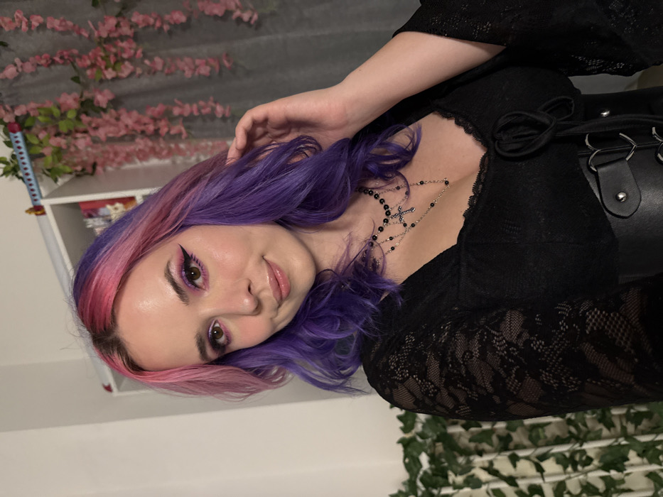 A young adult female, identified as Caucasian, is shown from the chest up in an indoor setting within the City of Zagreb, Croatia. She has wavy, split-dyed hair, transitioning from pink on the right (viewer's left) to purple on the left (viewer's right). Her facial features include brown eyes with prominent purple eyeshadow and black winged eyeliner, defined eyebrows, and a light pink lip gloss. She has a faint smile and is looking directly at the camera.
She is wearing a black, long-sleeved top with a visible lace pattern on the sleeves. Over this, a black faux leather corset-style garment with silver-toned buckles and studs is visible around her midsection. Around her neck, she wears multiple layered necklaces, including a black beaded rosary-style necklace with a silver crucifix pendant. Her left hand is raised, with fingers lightly touching the purple section of her hair near her ear.
The background consists of a light-colored wall. To the upper right, a white shelving unit is visible, upon which a small red object, possibly a katana hilt, rests horizontally. Artificial pink cherry blossom branches are draped over a dark background panel to the right, and artificial green ivy climbs vertically on a white lattice structure or similar support to the left. The scene is illuminated by soft, frontal lighting.