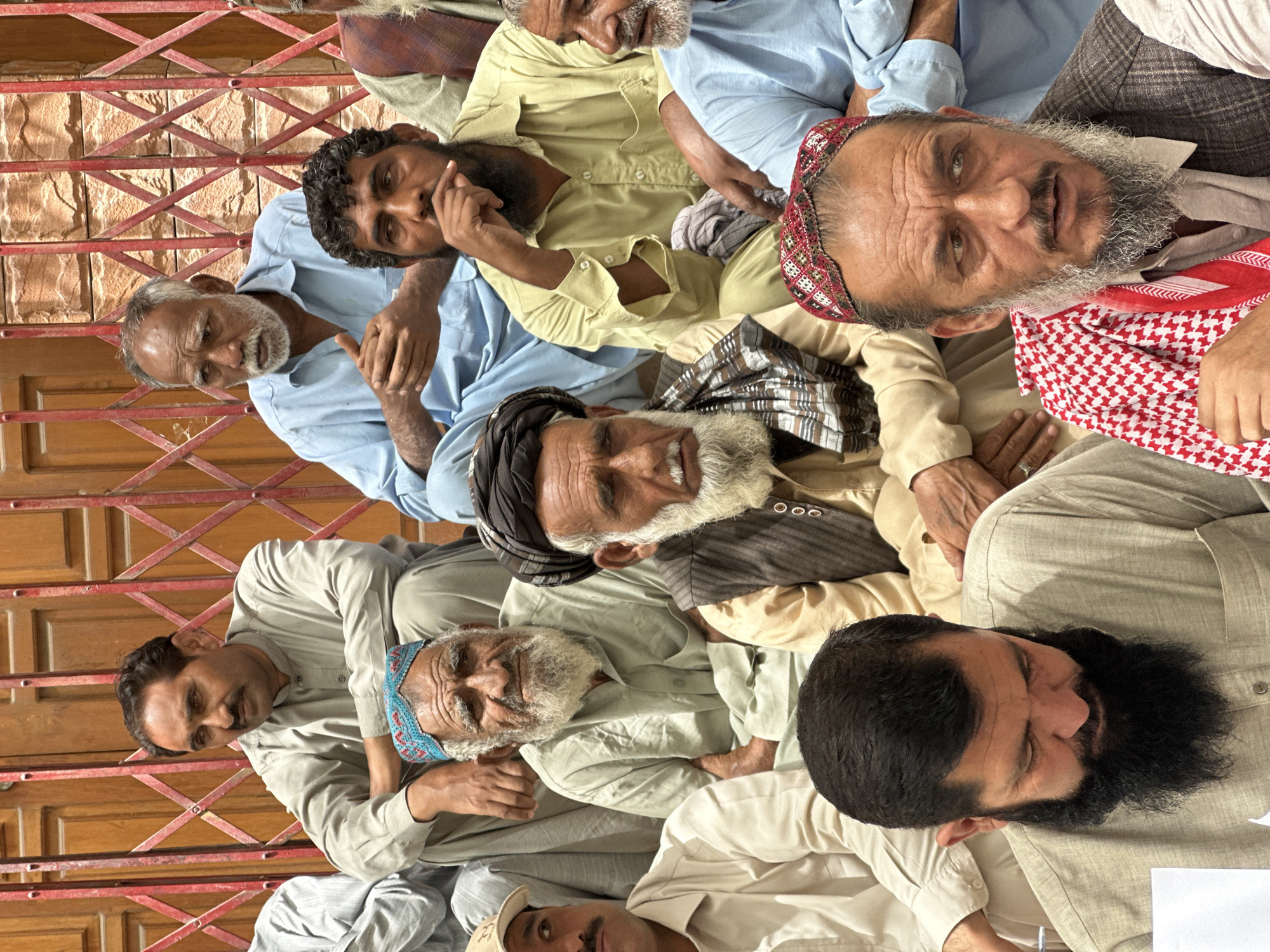 A group of men of varying ages are seated in front of a red metal gate, which appears to be a security barrier, in an outdoor setting. The background wall is made of rough-hewn stone. The men are dressed in traditional South Asian clothing, primarily shalwar kameez. Some are wearing caps or turbans. They appear to be gathered for a meeting or event, with some looking directly at the camera and others looking away. The overall atmosphere is informal. This scene is located in Quetta City Tehsil, Pakistan.