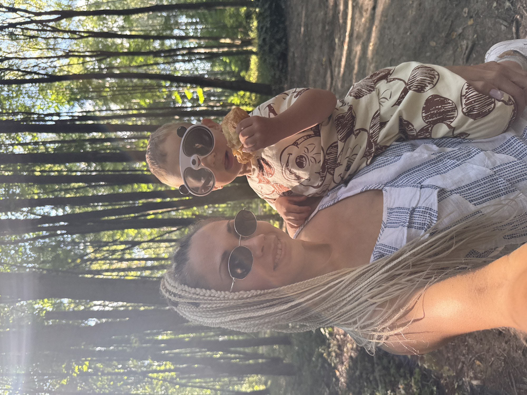 In an outdoor setting resembling a sun-dappled forest, likely in the city of Dumino, Russia, a woman with blonde braided hair is holding a young child. The woman is wearing sunglasses and a striped top. The child, also wearing sunglasses designed to look like a panda's face, is holding and eating a pastry. The background is filled with tall trees and vegetation, suggesting a park or wooded area. The lighting indicates it is daytime, with sunlight filtering through the leaves.