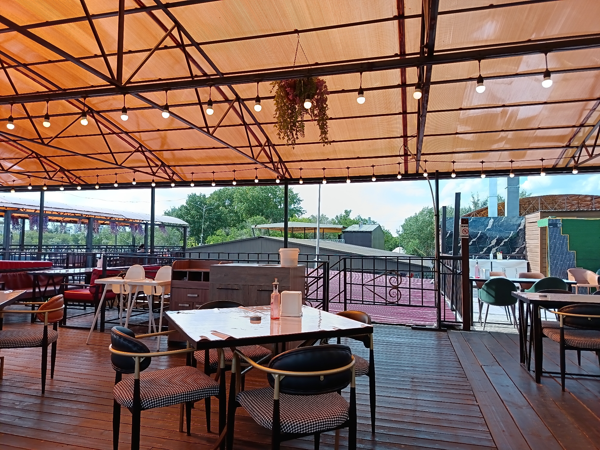The image depicts an outdoor seating area of a restaurant or cafe, likely located in Qarağandı city administration, Kazakhstan. The scene is characterized by a large, orange, translucent canopy supported by a dark metal framework, under which several tables and chairs are arranged. String lights are suspended from the canopy, and a hanging plant arrangement is visible in the center.
In the foreground, several dining tables with chairs are set up on a wooden deck. One table is covered with a clear plastic sheet, and on it are a pink bottle and a white container. The chairs have dark frames and patterned upholstery. Further back, other tables and seating arrangements are visible, including red benches and white high chairs.
Beyond the immediate seating area, a black metal railing with decorative scrollwork separates the deck from a lower level, which also appears to have seating and a red-colored roof. In the background, there are trees, various buildings, and what appears to be industrial elements such as chimneys or ventilation systems. The sky is visible and appears to be partly cloudy. No people are clearly visible or identifiable in the image, though there might be indistinct figures in the background. The overall impression is that of a contemporary, open-air dining establishment.